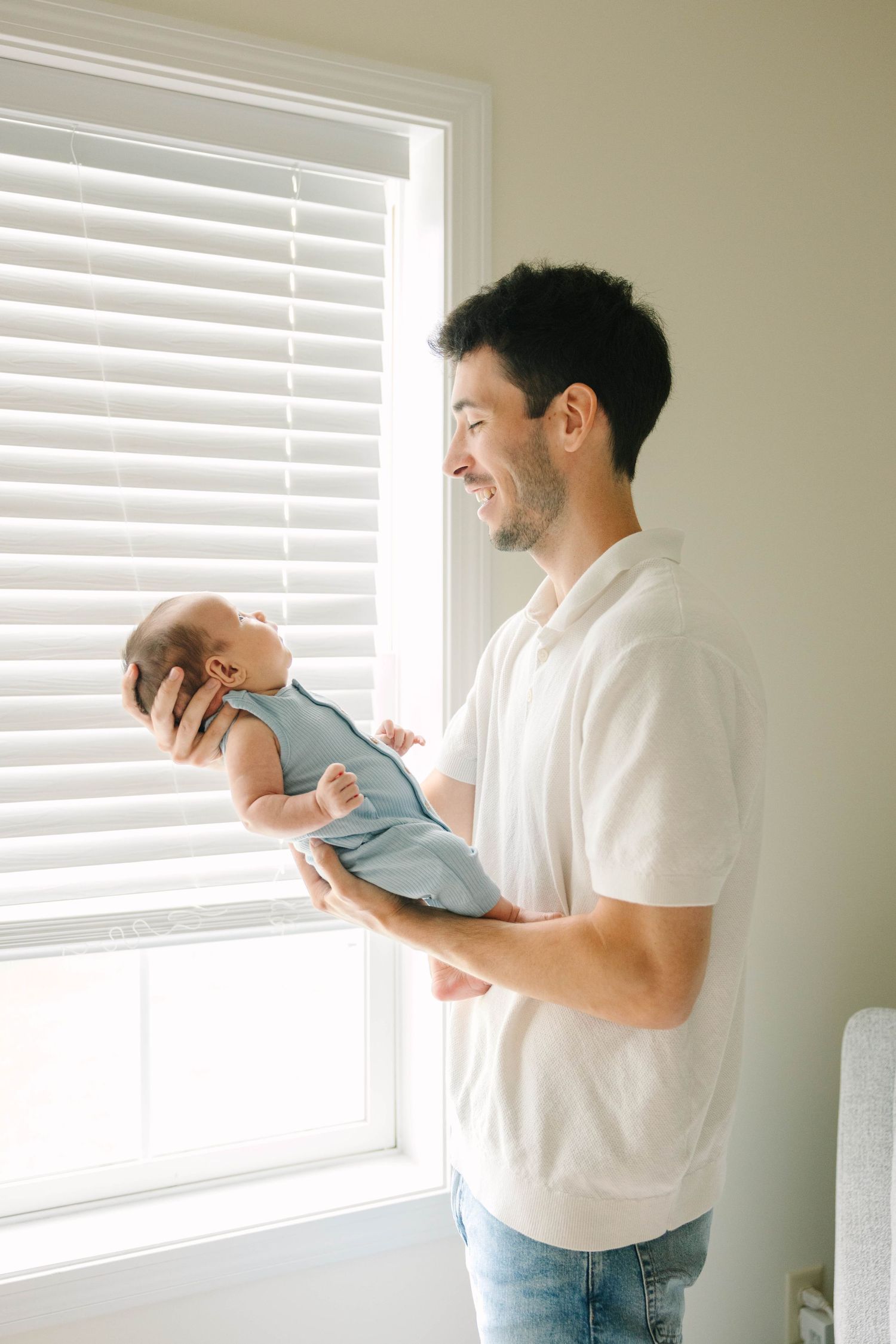 Smiling father holding his newborn baby in a blue romper near a bright window.