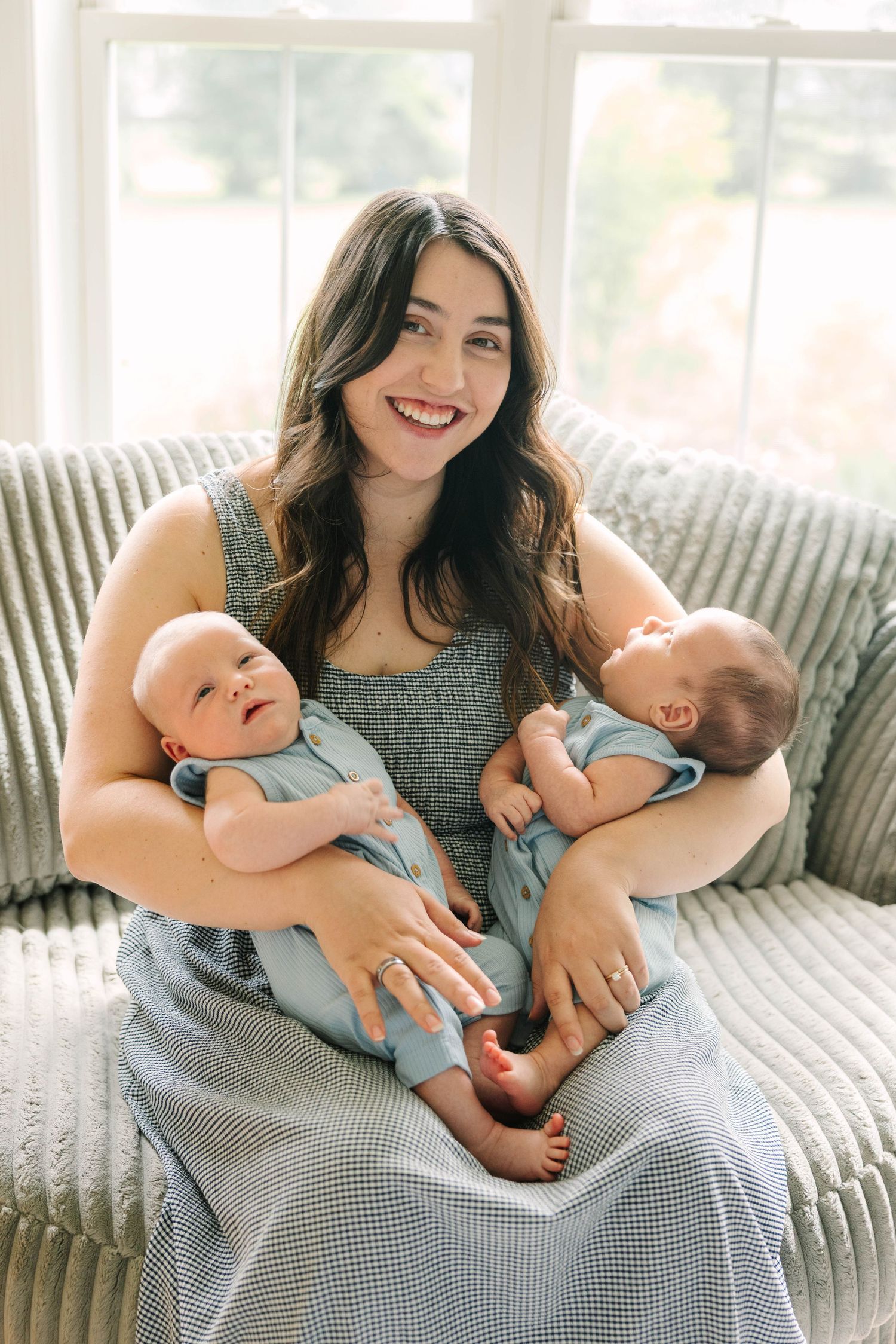 Smiling brunette mother in blue dress holding twin newborn babies on a textured gray sofa.