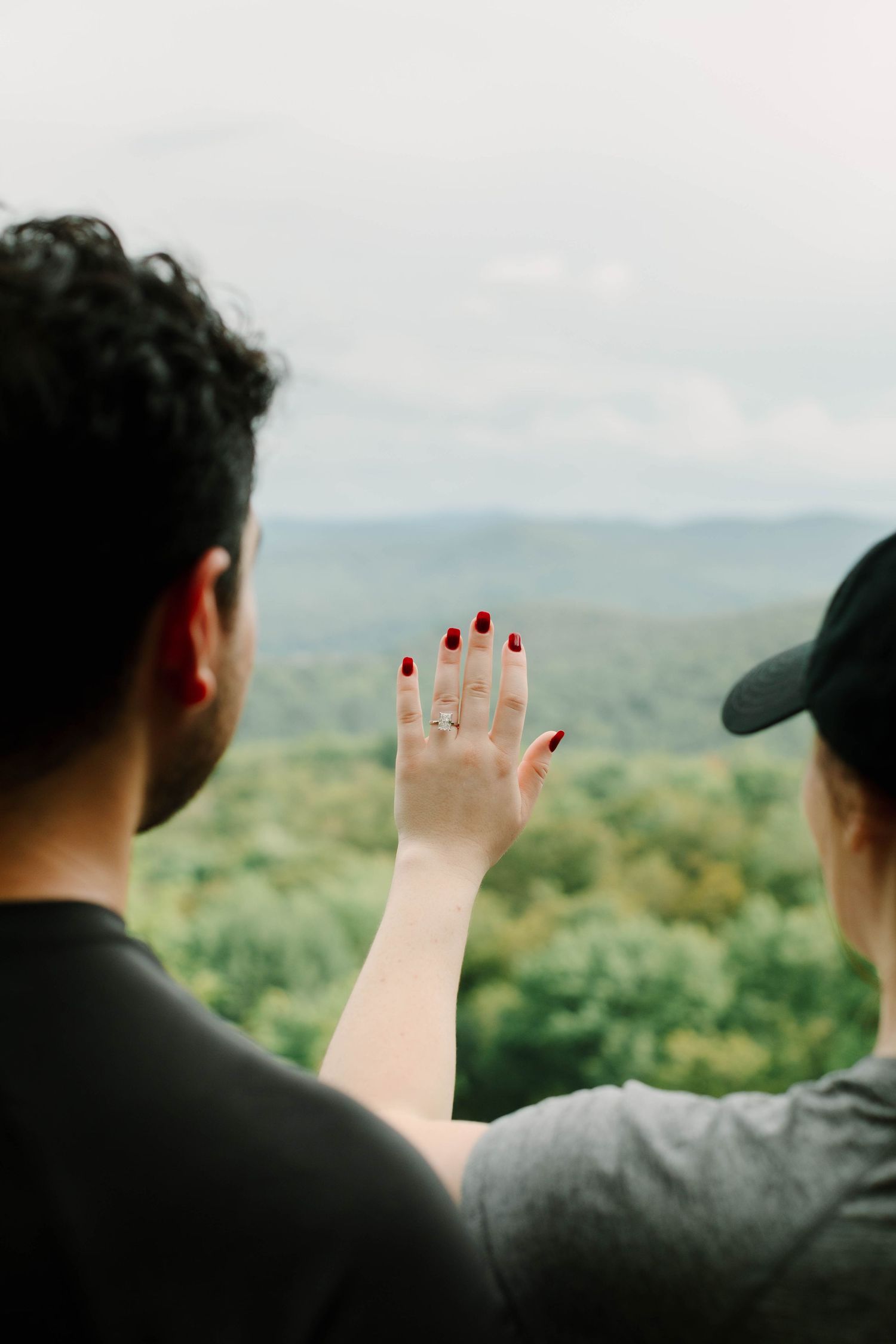 Couple admires new diamond engagement ring as woman holds hand up against scenic green mountain landscape.