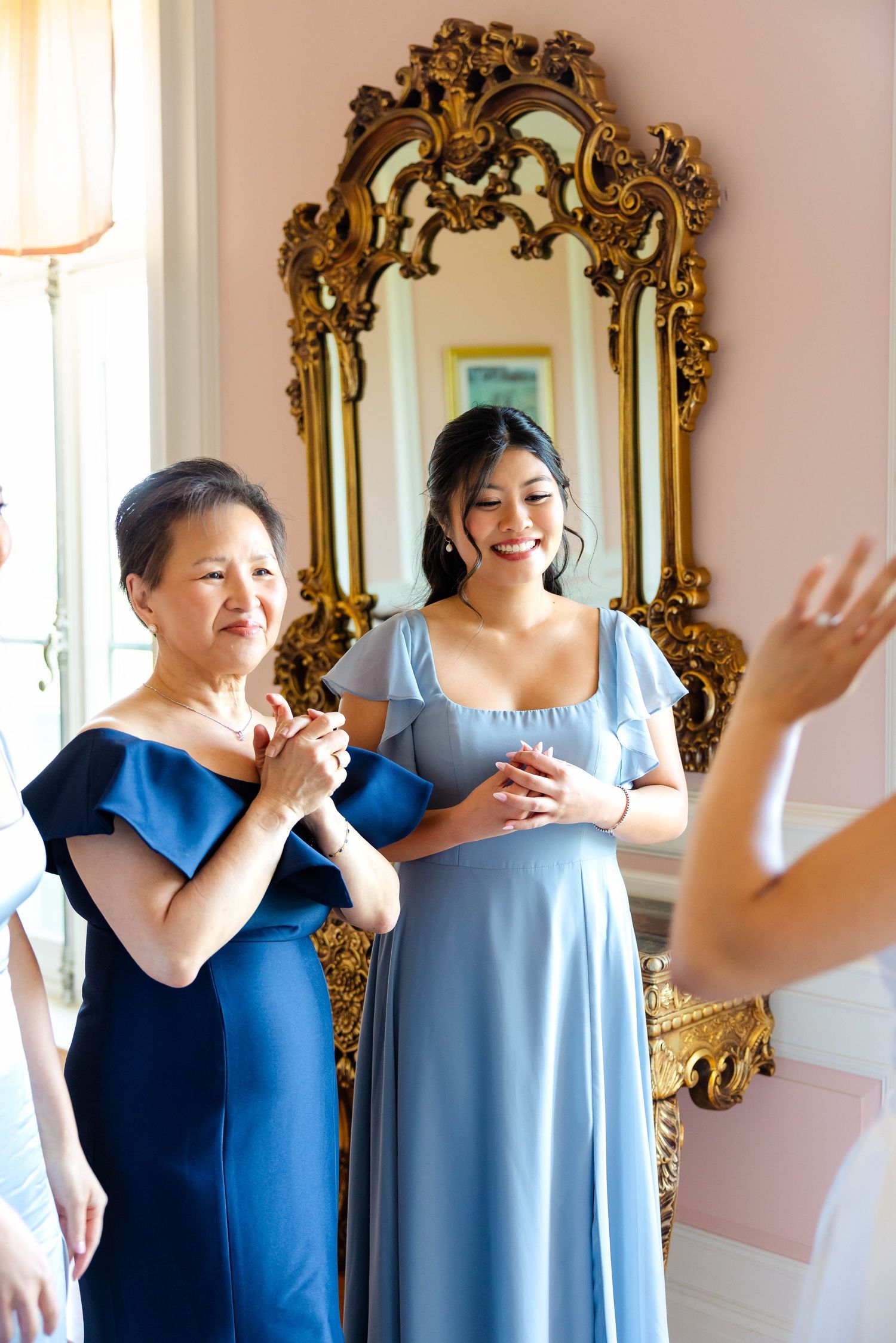 Two women in blue dresses smile joyfully before a ornate gold mirror during wedding preparations.