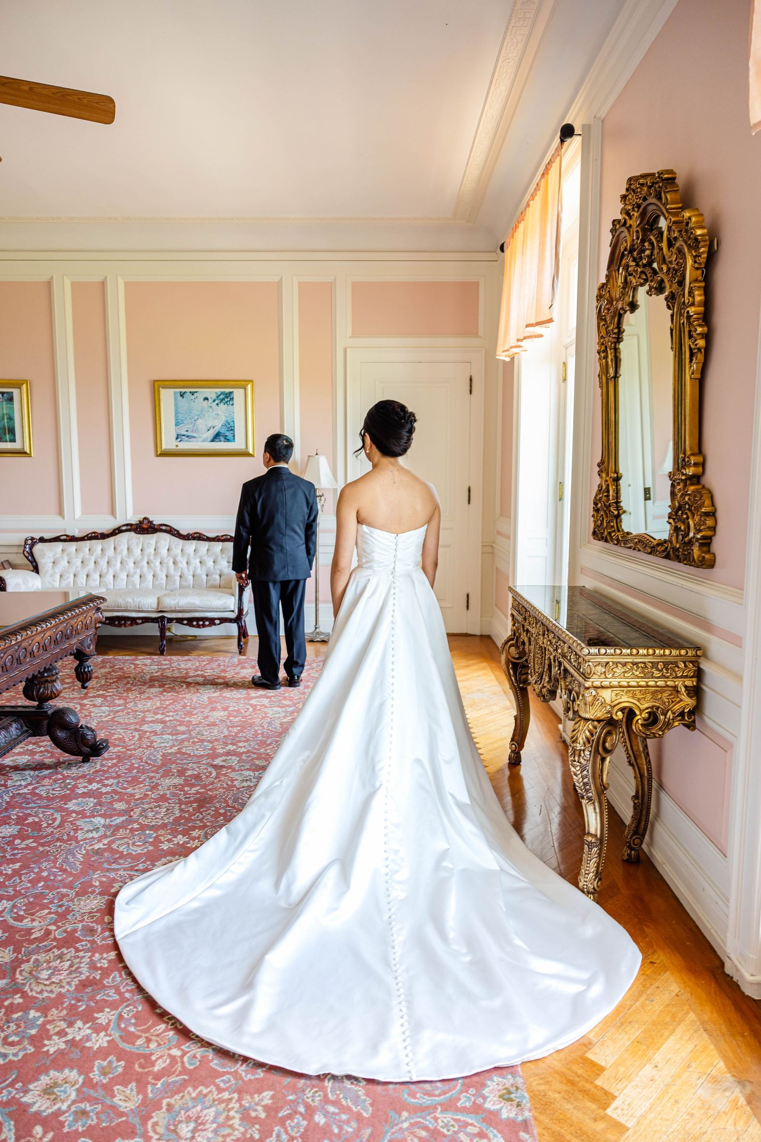 Bride in a strapless ballgown with cathedral train approaches groom for first look in ornate pink room.
