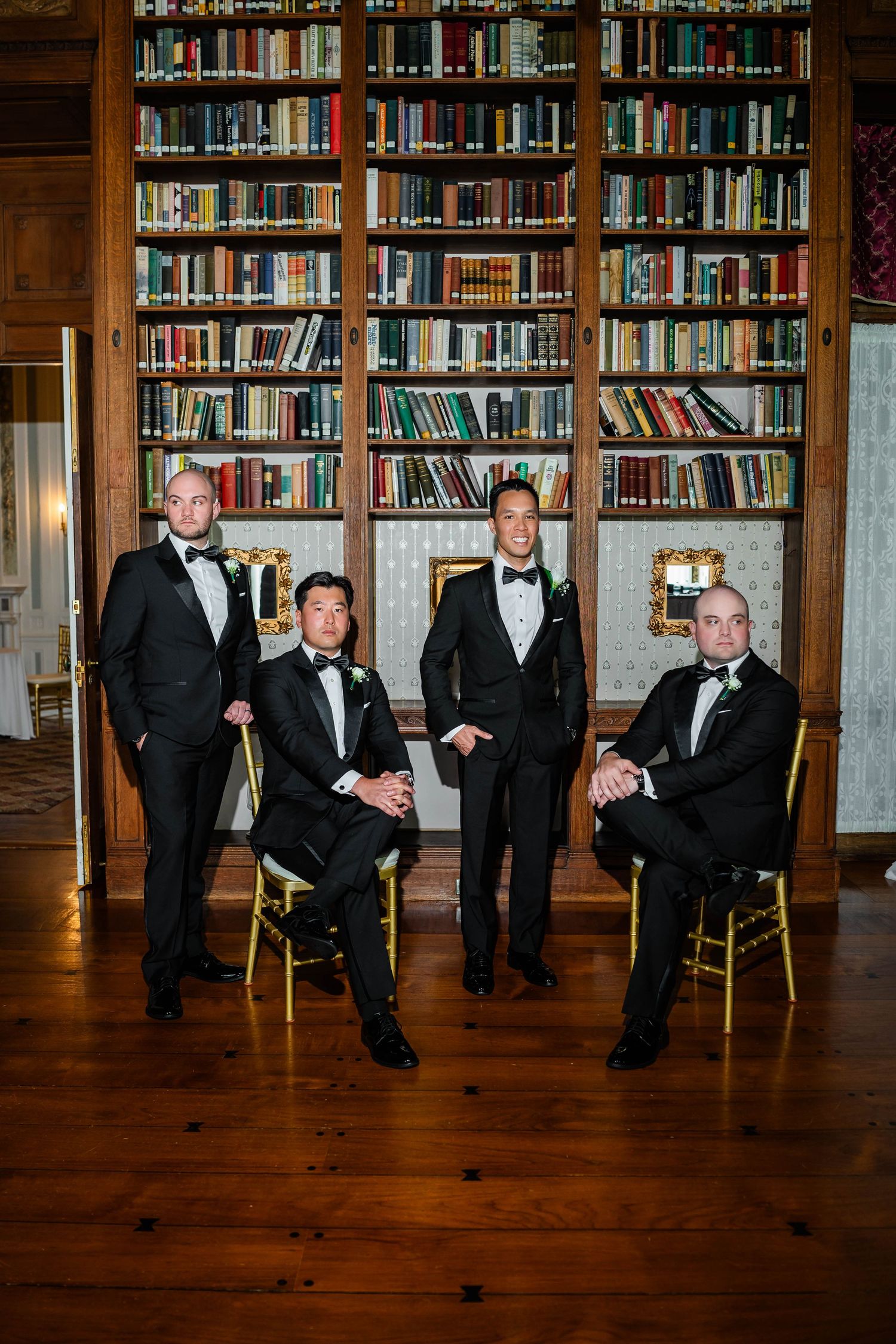 Four groomsmen in black tuxedos posing in front of floor-to-ceiling bookshelves in library.