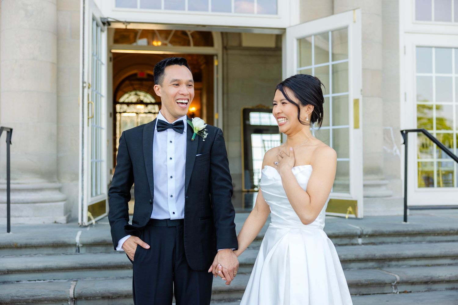 Happy bride and groom in formal wedding attire laughing together on steps outside a grand building.
