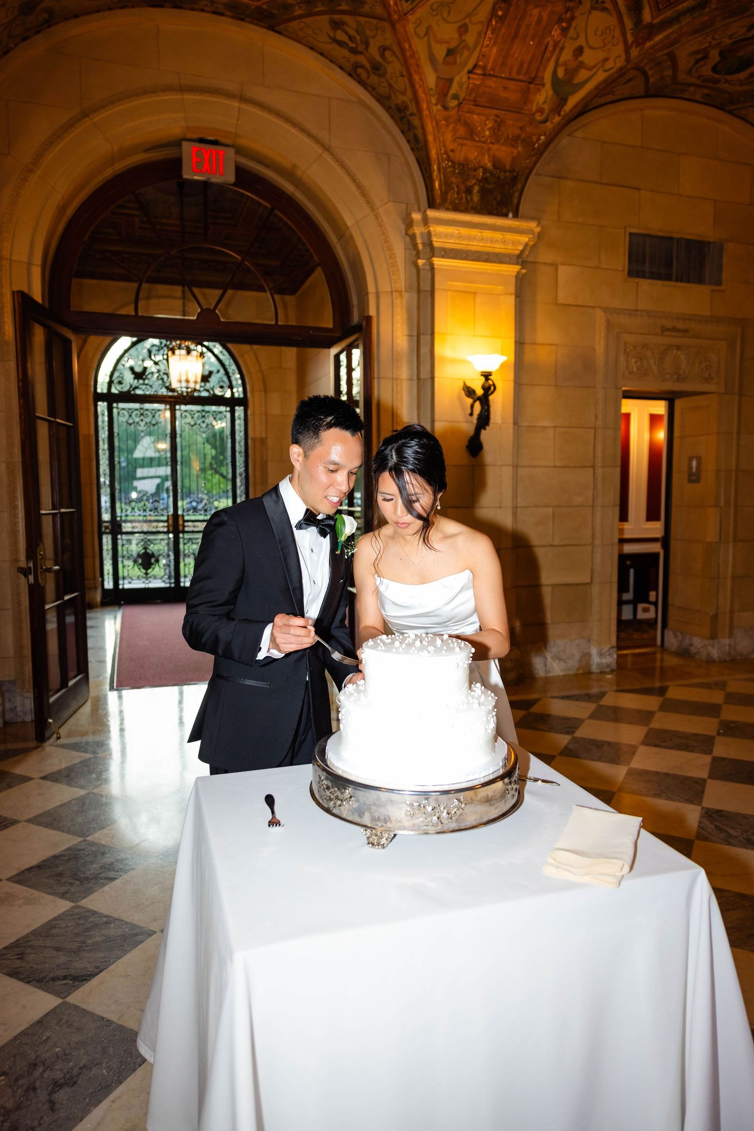 Asian bride and groom cut white tiered wedding cake in ornate marble hall with arched doorways and painted ceiling.