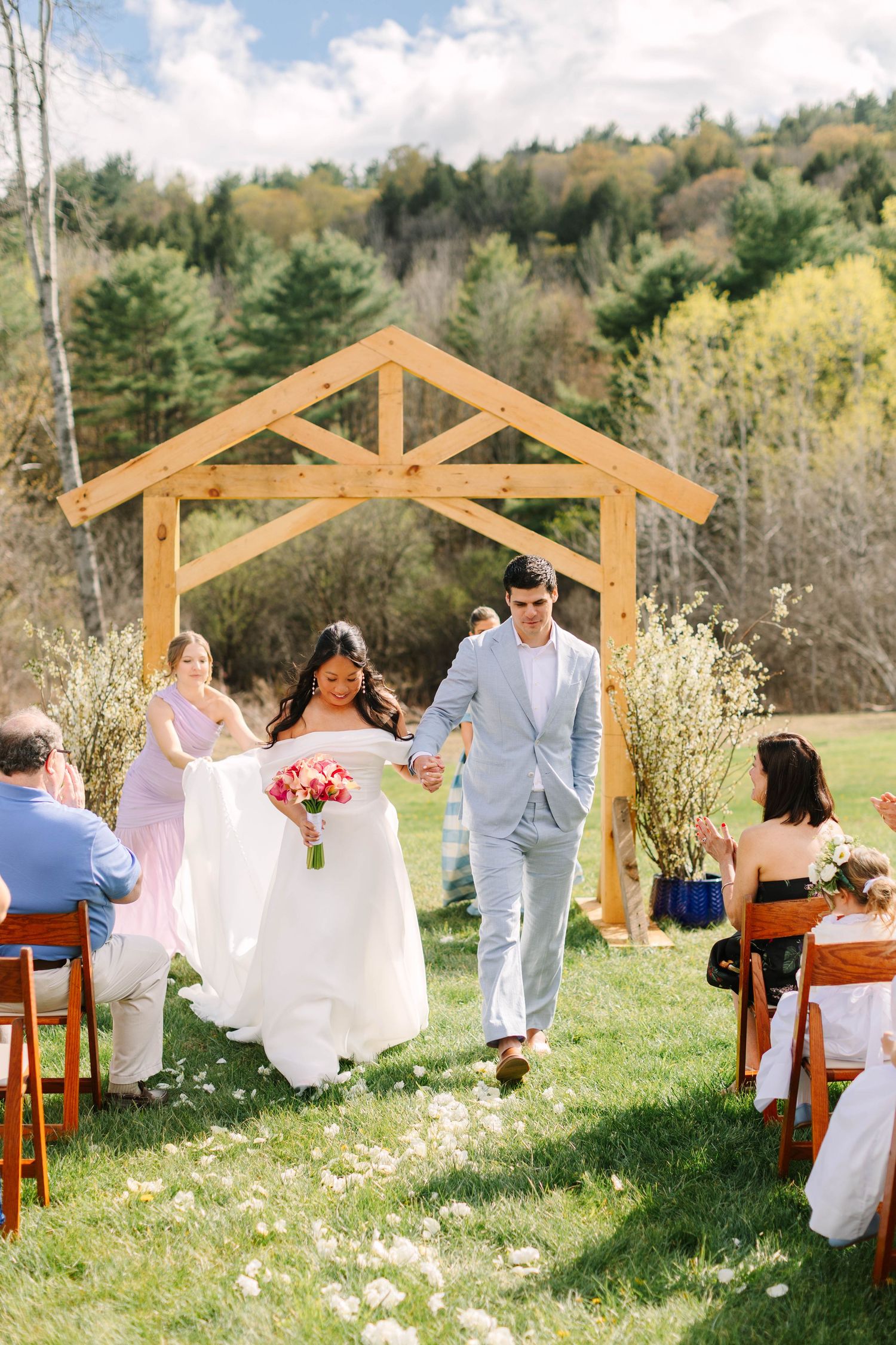 Newlyweds walk down petal-lined aisle under wooden arch at outdoor ceremony with forest backdrop.