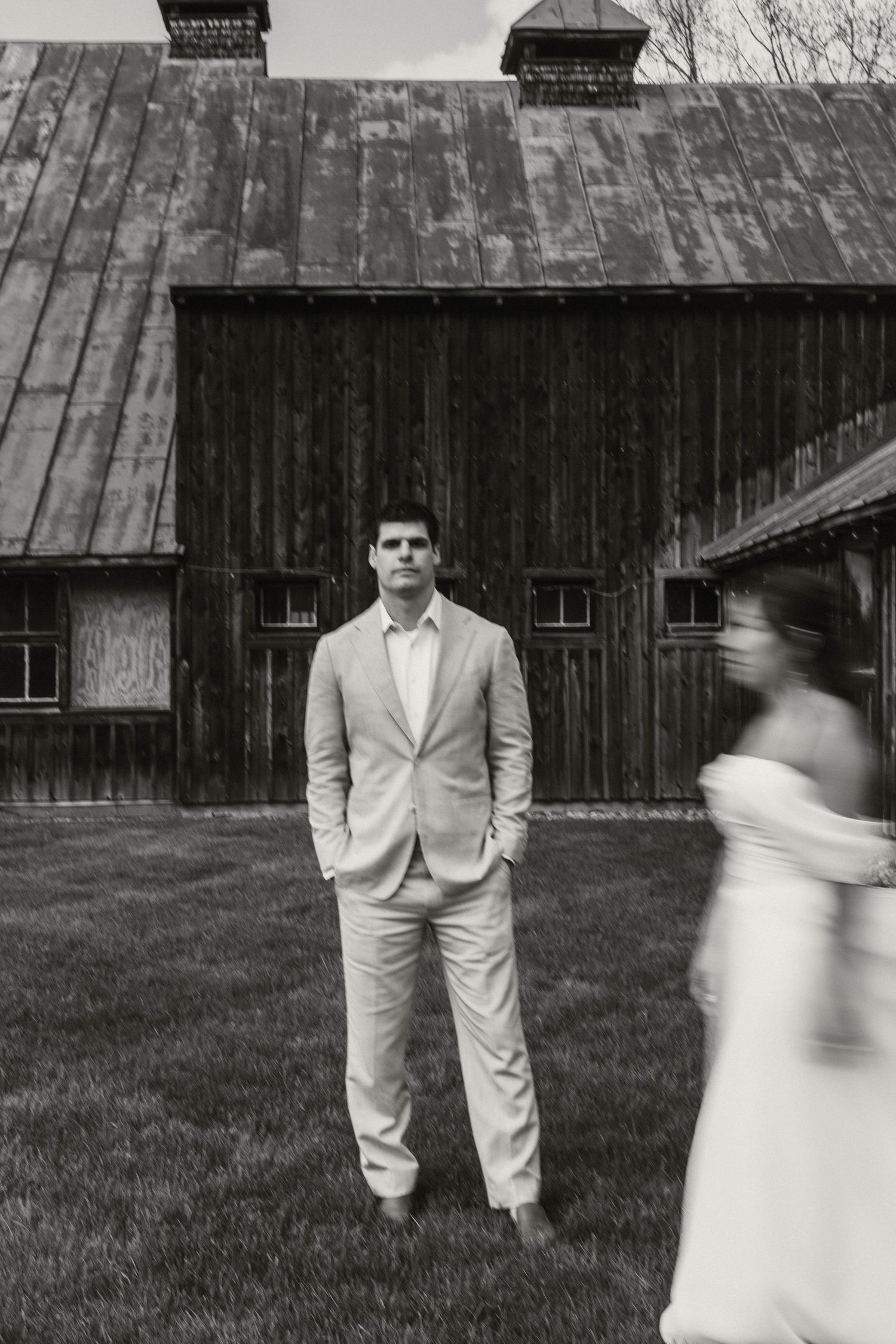 Black and white photo of groom standing before rustic barn as bride approaches in blur.