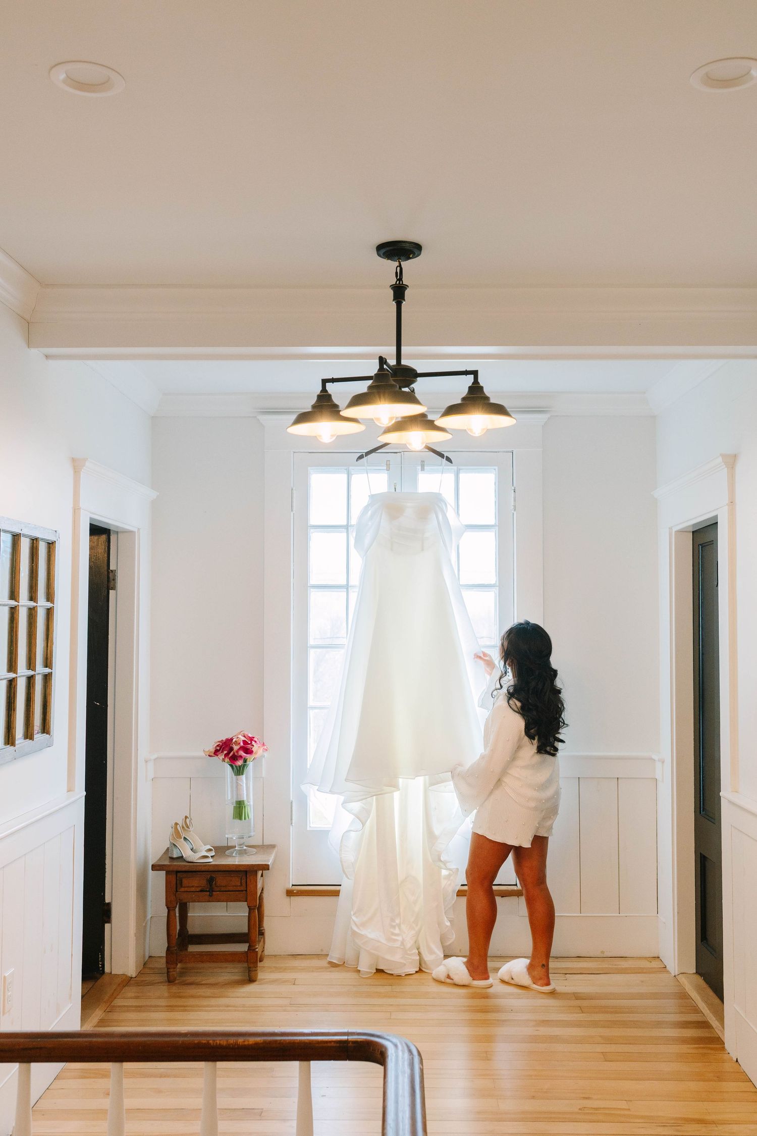 Bride in white robe reaching up to touch her elegant wedding gown hanging by a window in a bright room.