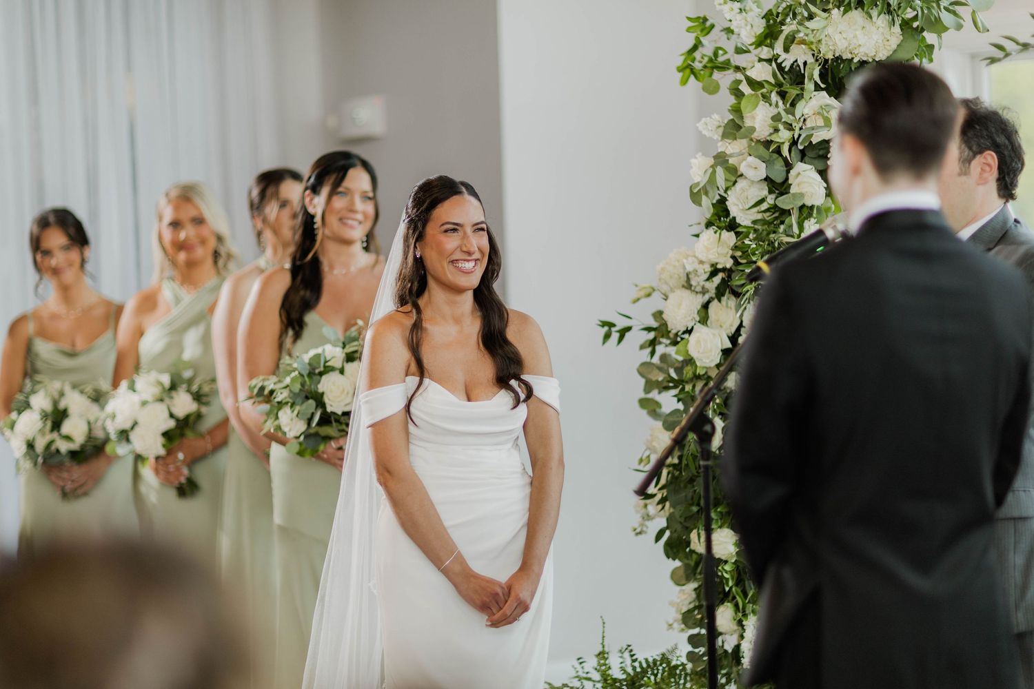 Smiling bride in off-shoulder white gown stands at altar with bridesmaids in sage green dresses behind her.