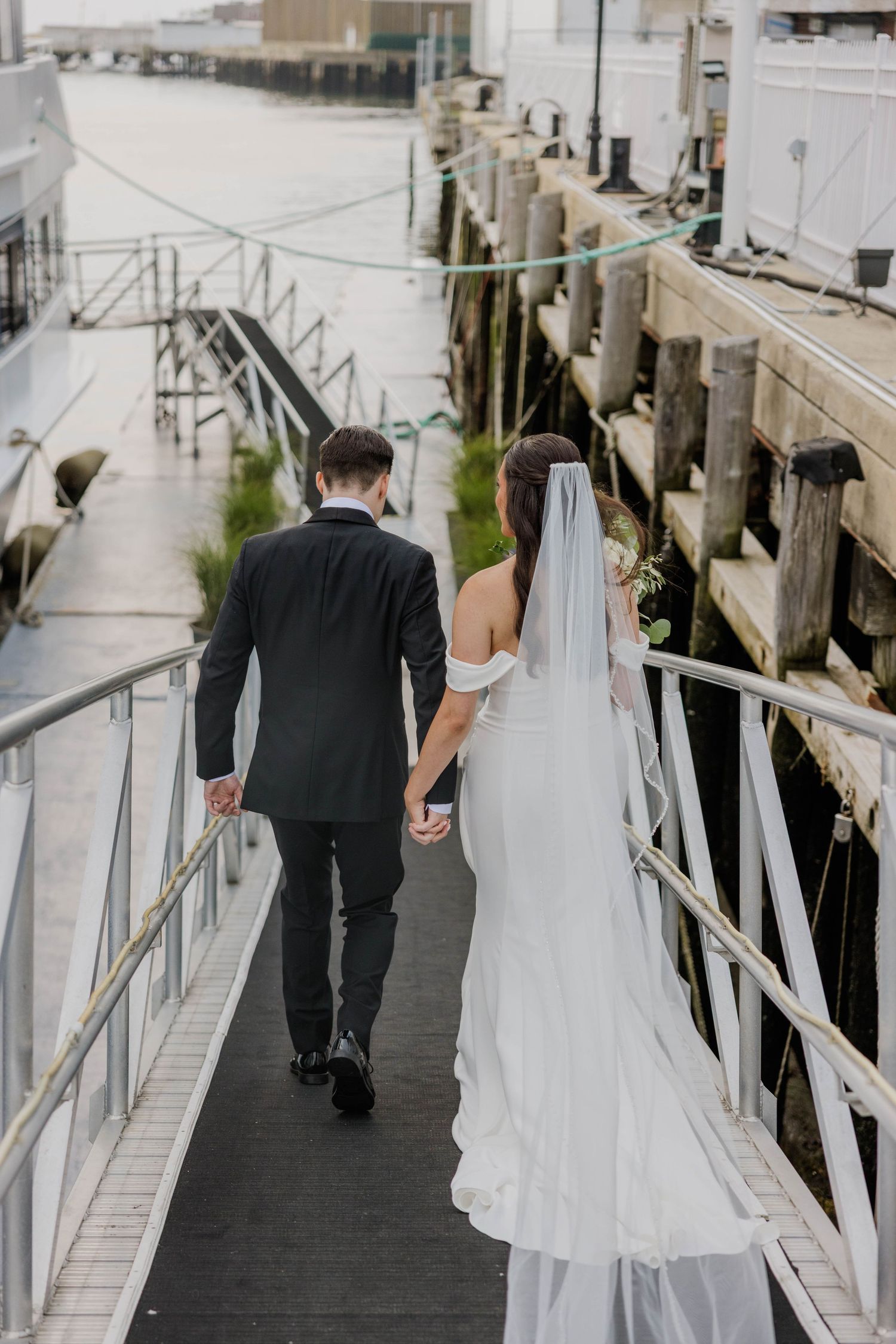 Bride and groom walk hand in hand along a waterfront dock after their wedding ceremony.