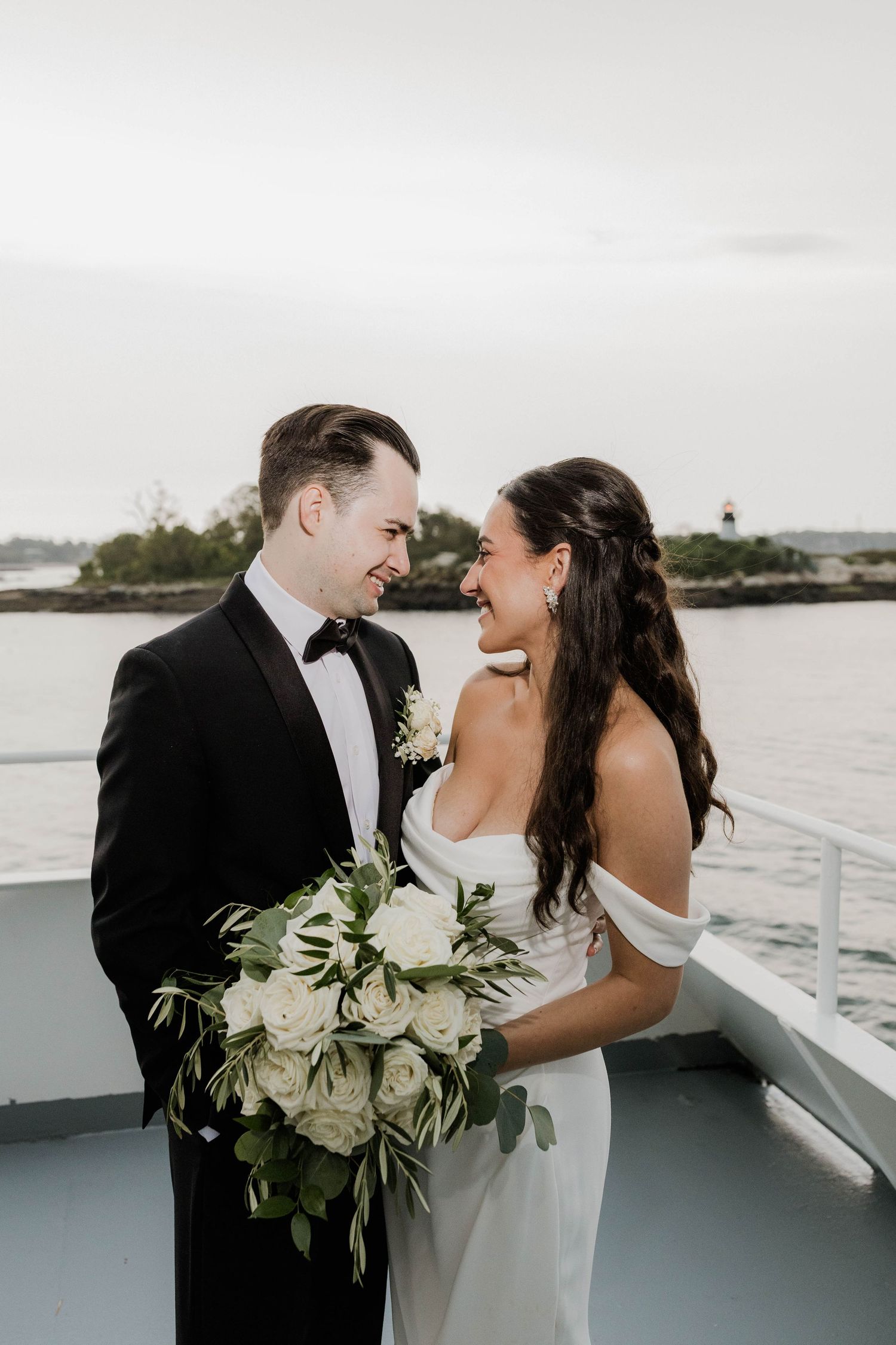 Bride in off-shoulder white gown and groom in tuxedo share a laugh on boat deck by water.