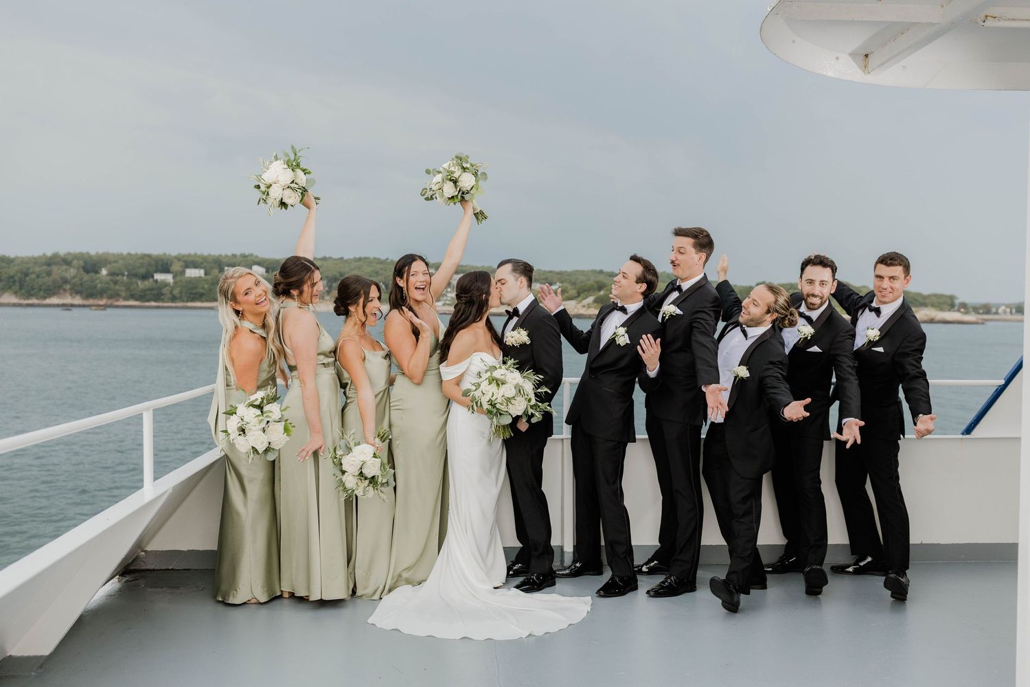 Wedding party celebrates on boat deck, bridesmaids in sage green dresses with groomsmen in black tuxedos.