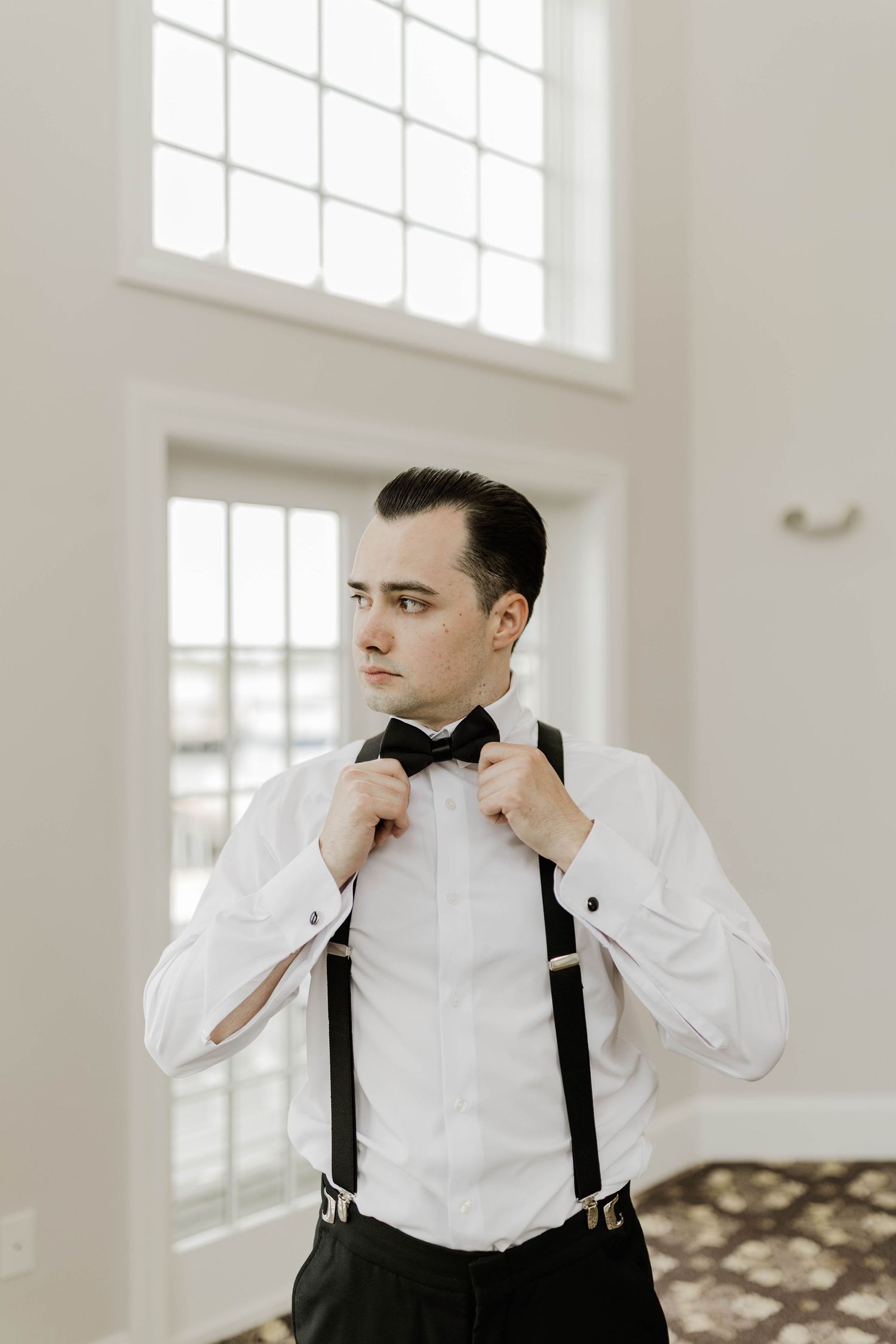 Groom in white dress shirt adjusting black bow tie with suspenders, standing near large window in bright room.