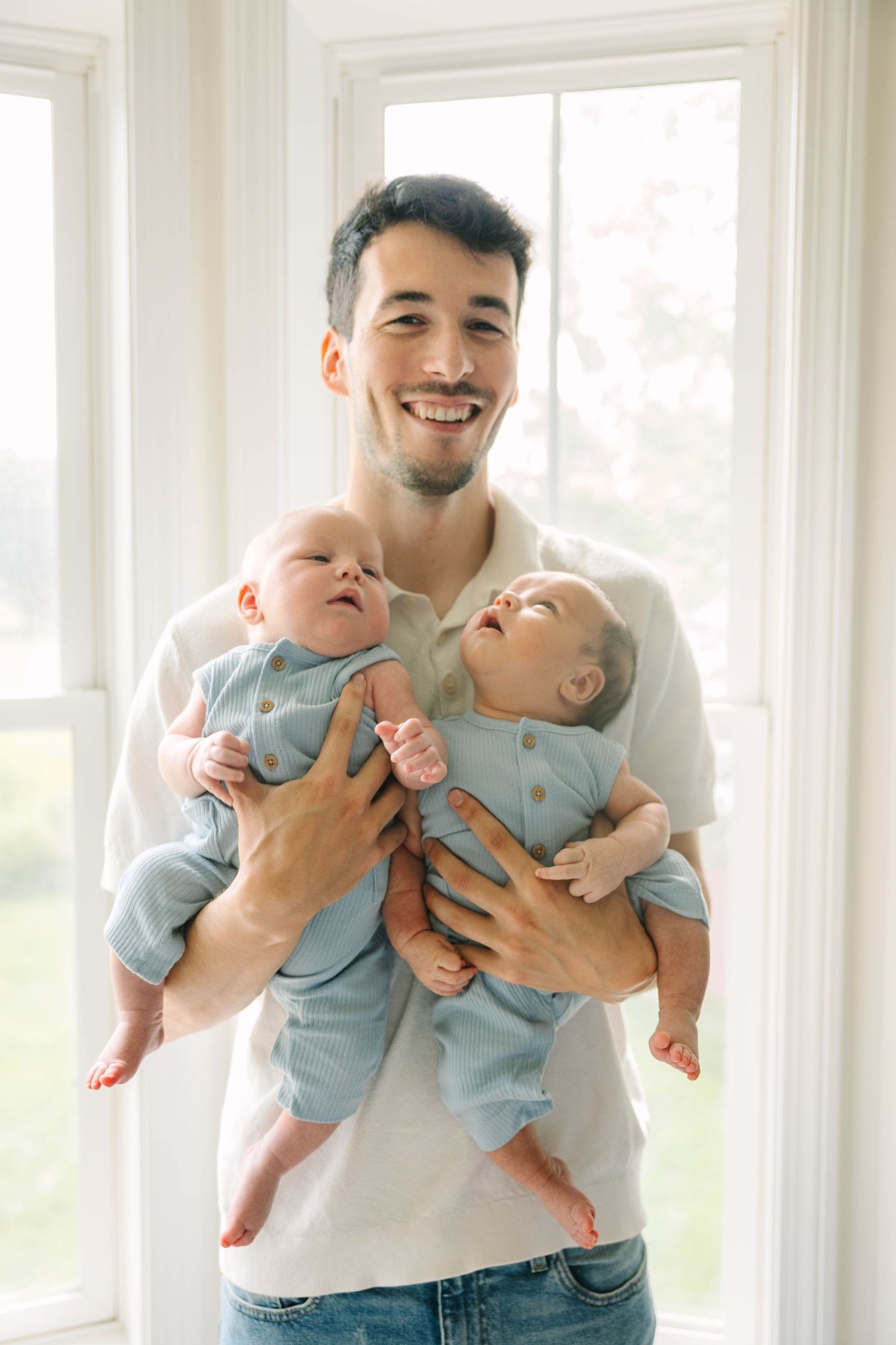 Proud father smiling while holding twin babies dressed in blue outfits by large windows.