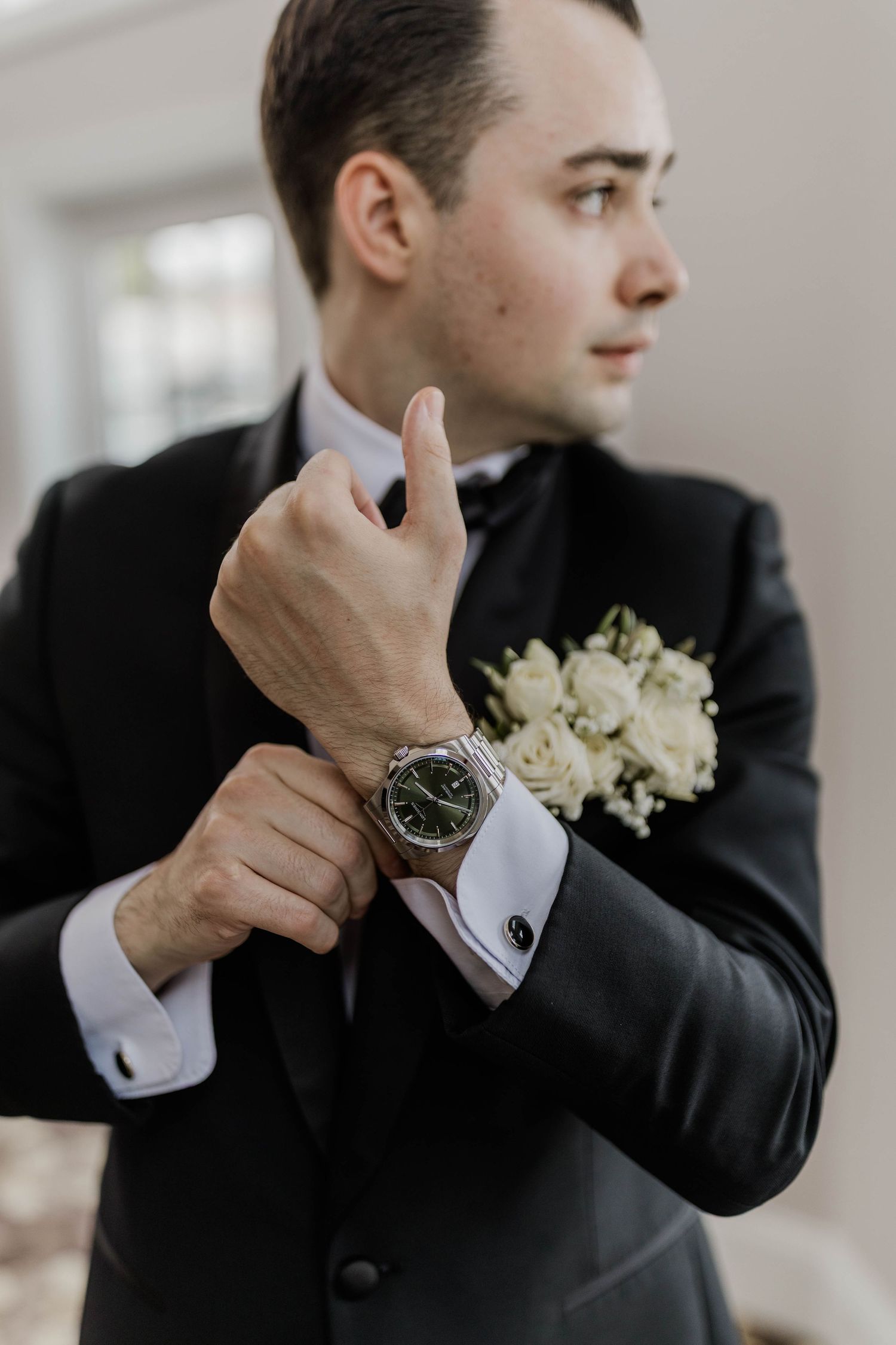 Groom in black tuxedo adjusting cufflinks with boutonniere and silver watch.