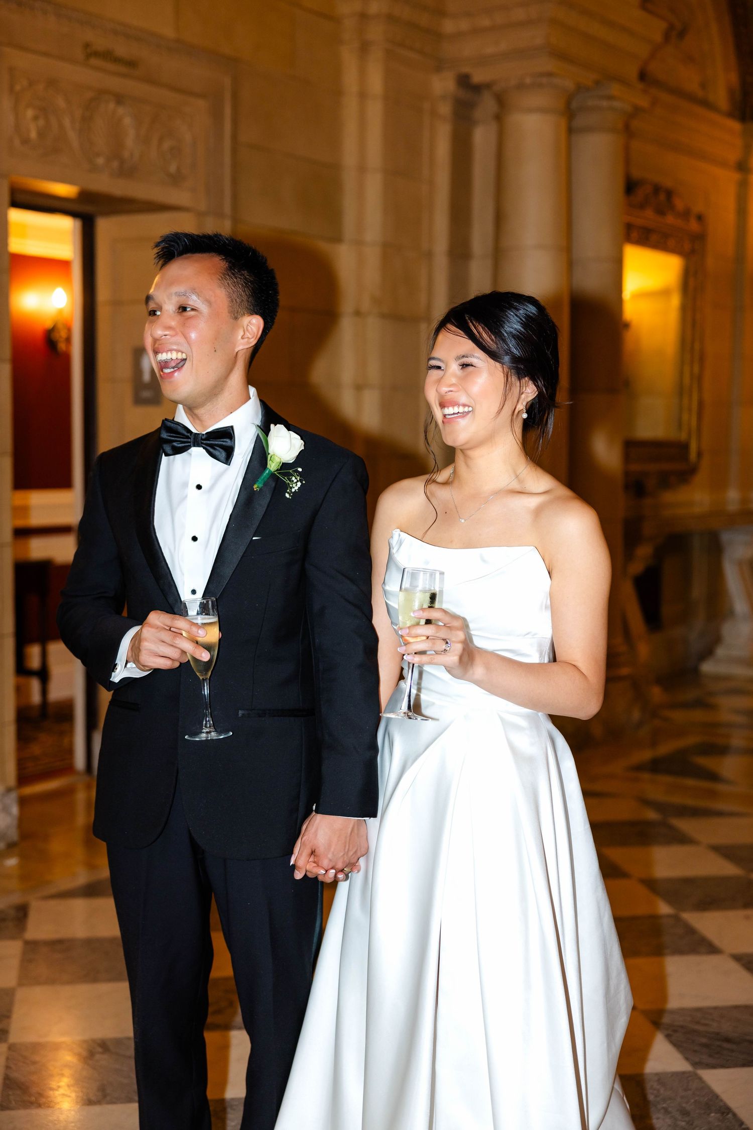 Smiling bride and groom holding champagne glasses hand in hand in ornate golden reception hall.
