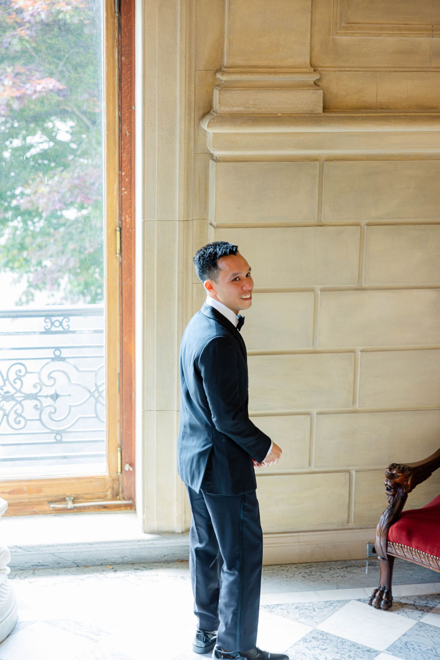 Elegant groom in black tuxedo stands by grand window in luxury venue, glancing over shoulder.