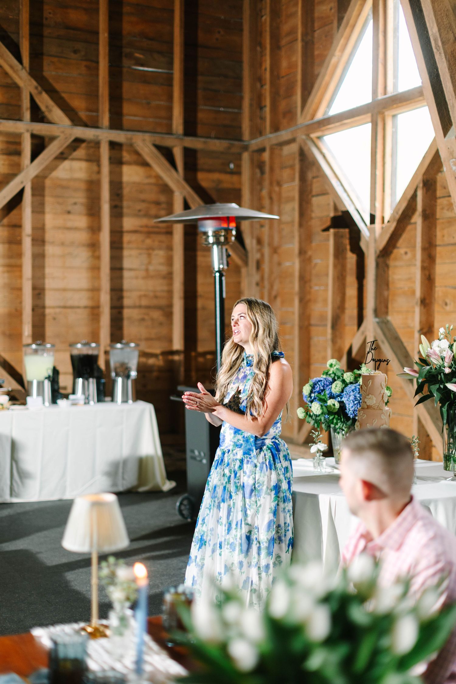 Woman in blue floral dress gives a speech inside a rustic barn wedding reception venue with wood beam ceiling.