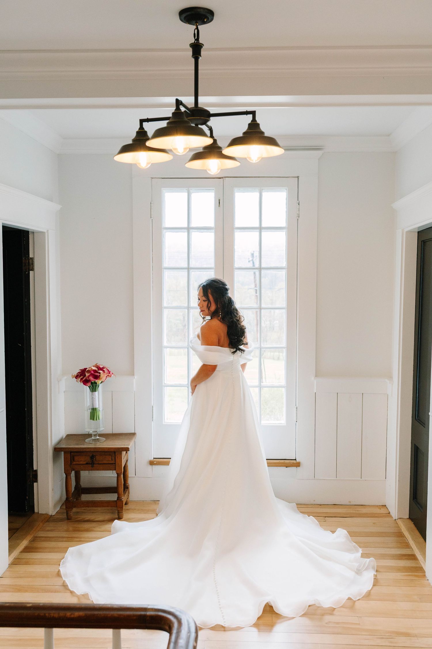 Bride in off-shoulder white gown with long train stands by window in beautifully lit room.