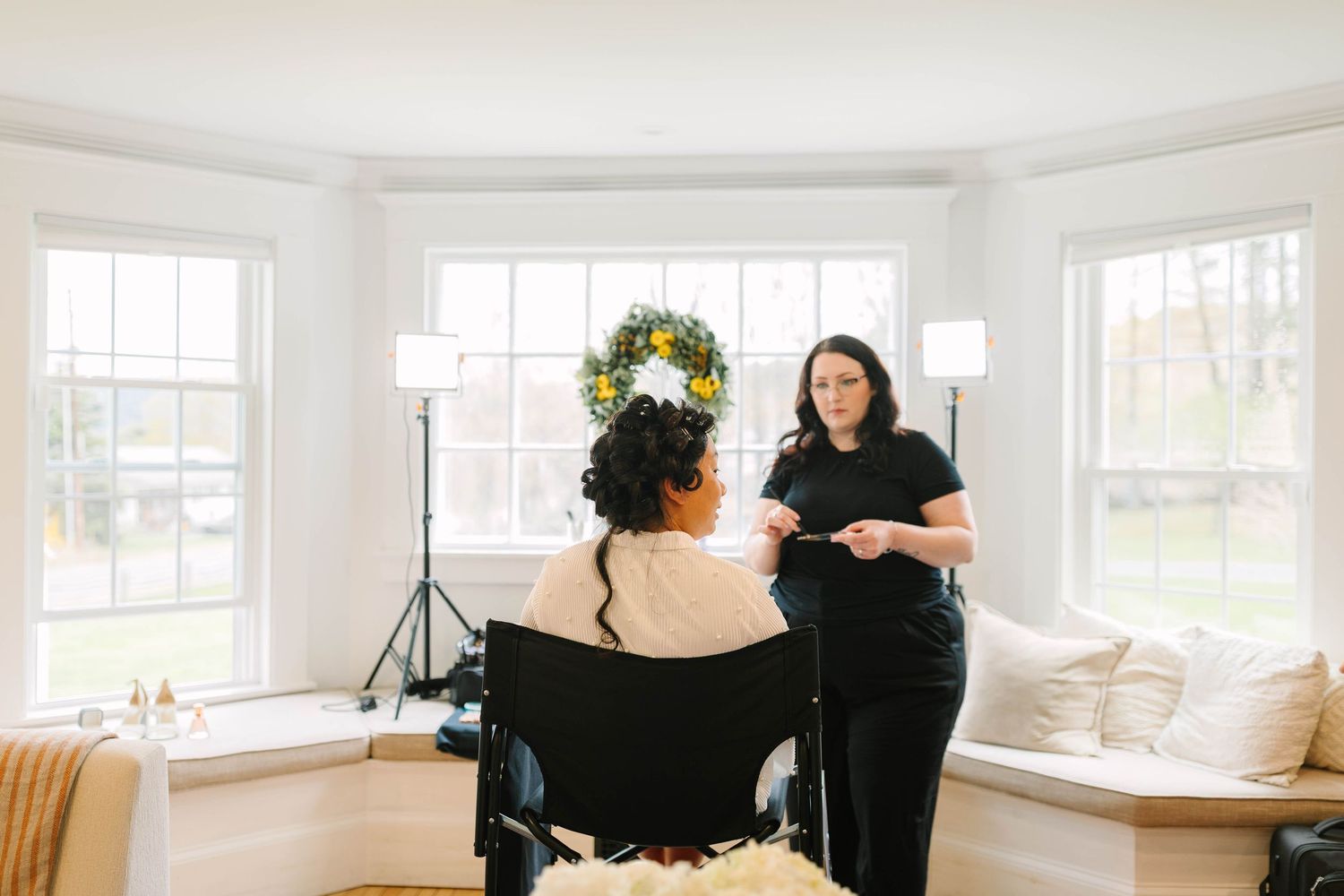 Bride seated in chair with floral crown while stylist prepares her in bright white room with large windows.