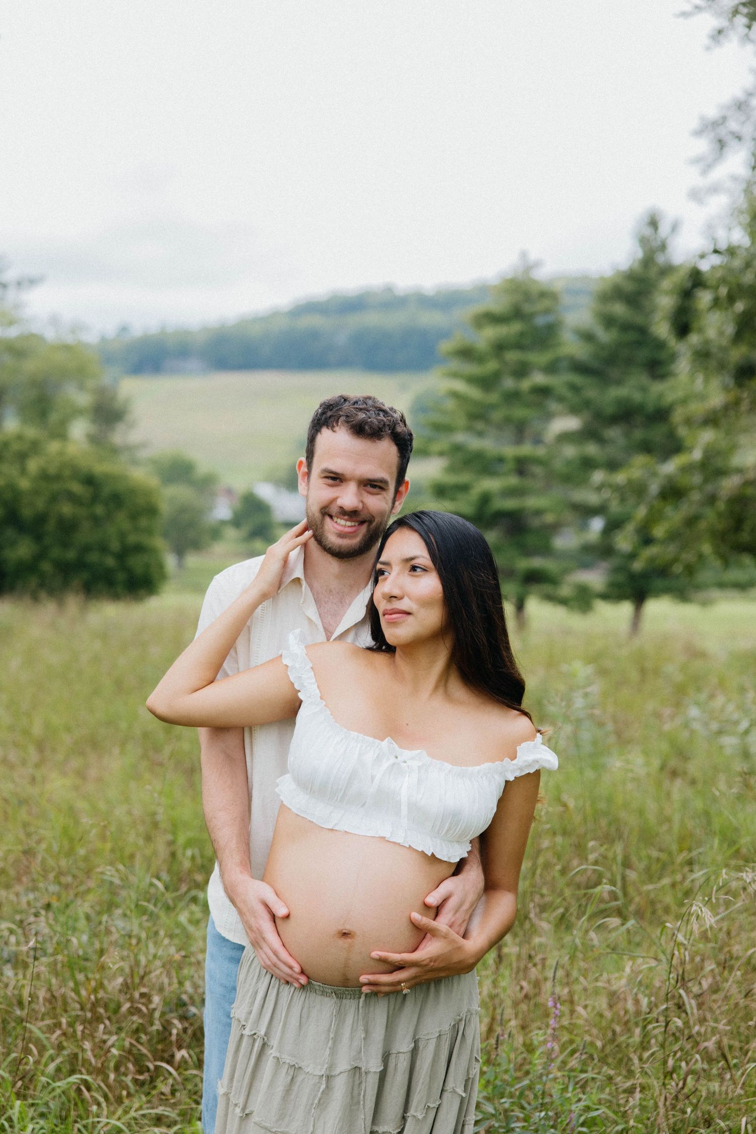 Happy couple poses in a green field, woman in white crop top showing baby bump, man embracing her from behind.
