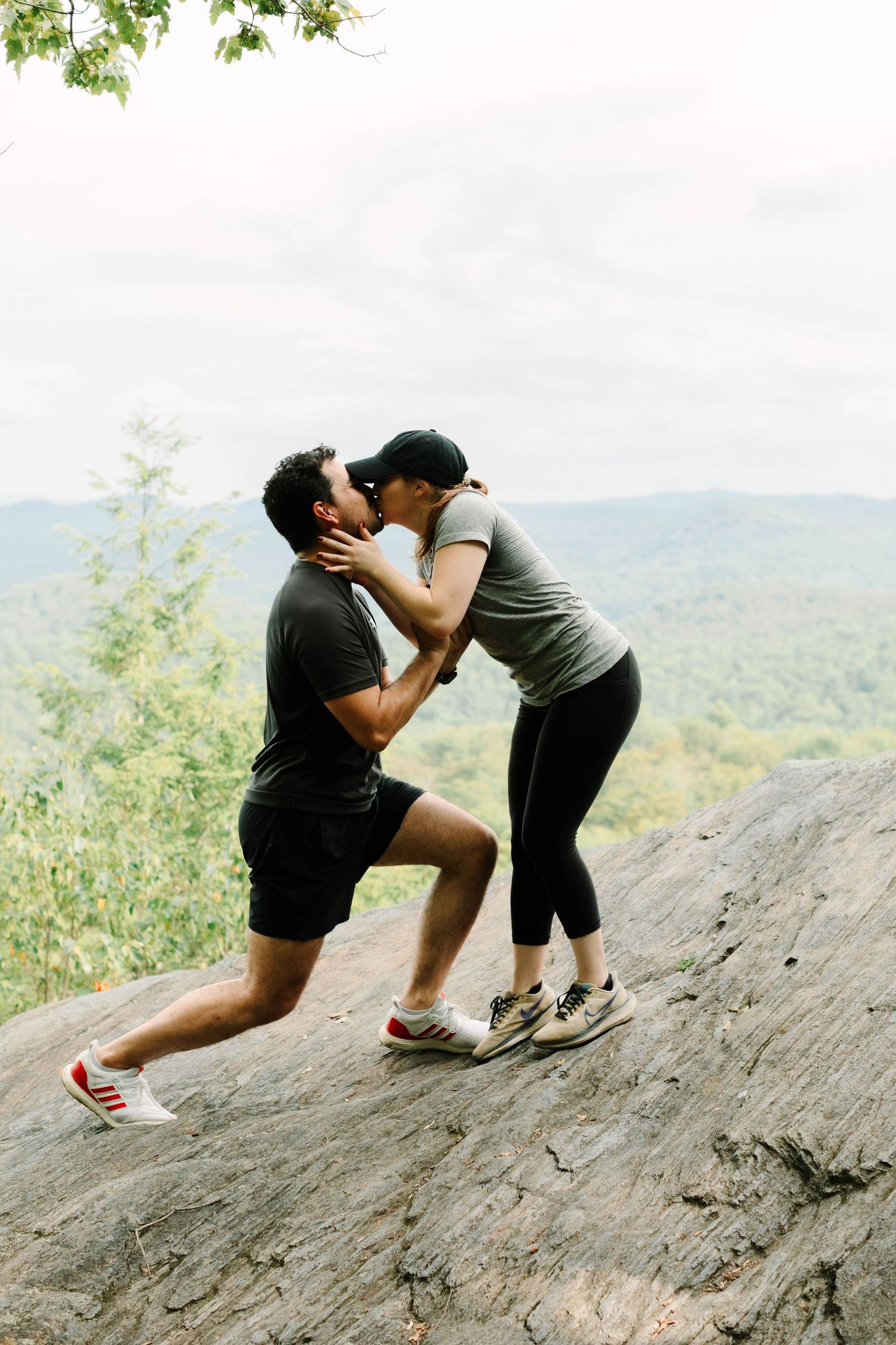 A couple shares a romantic kiss on a rocky overlook with lush green mountain views during an outdoor hiking proposal.