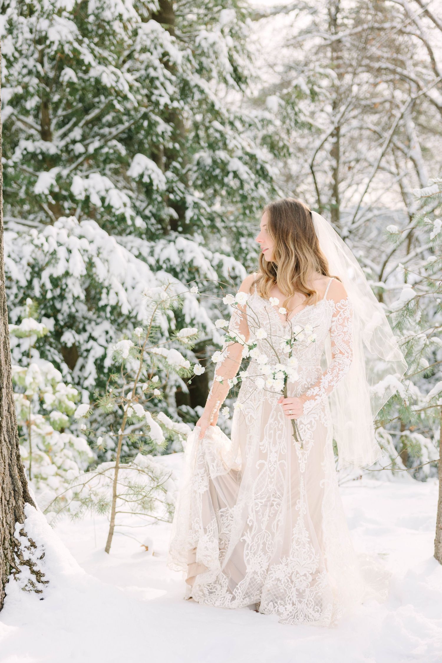 Elegant bride in lace wedding dress with veil walking through snow-covered pine forest.