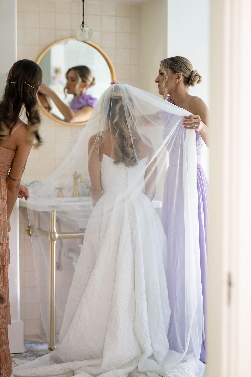 Bridesmaids helping bride with veil in elegant bathroom before wedding ceremony.