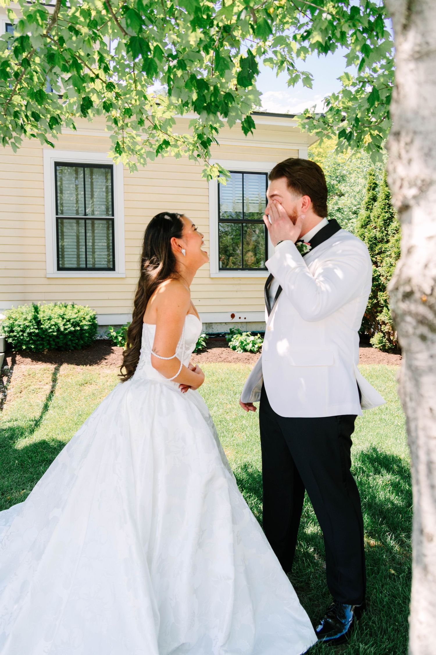 Groom in white tuxedo covering mouth in awe while bride in strapless gown laughs outdoors.