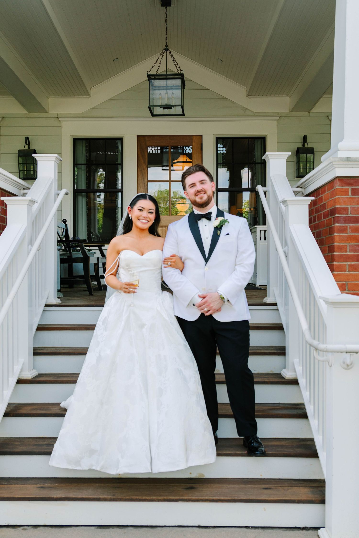 Bride and groom posing on white staircase of elegant venue, groom in white tuxedo jacket with black bow tie.