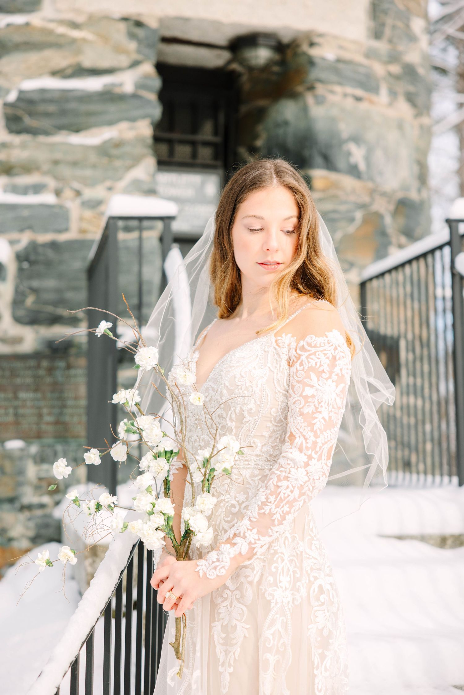Bride in lace gown and veil gazes down holding white floral bouquet near stone building in snow.