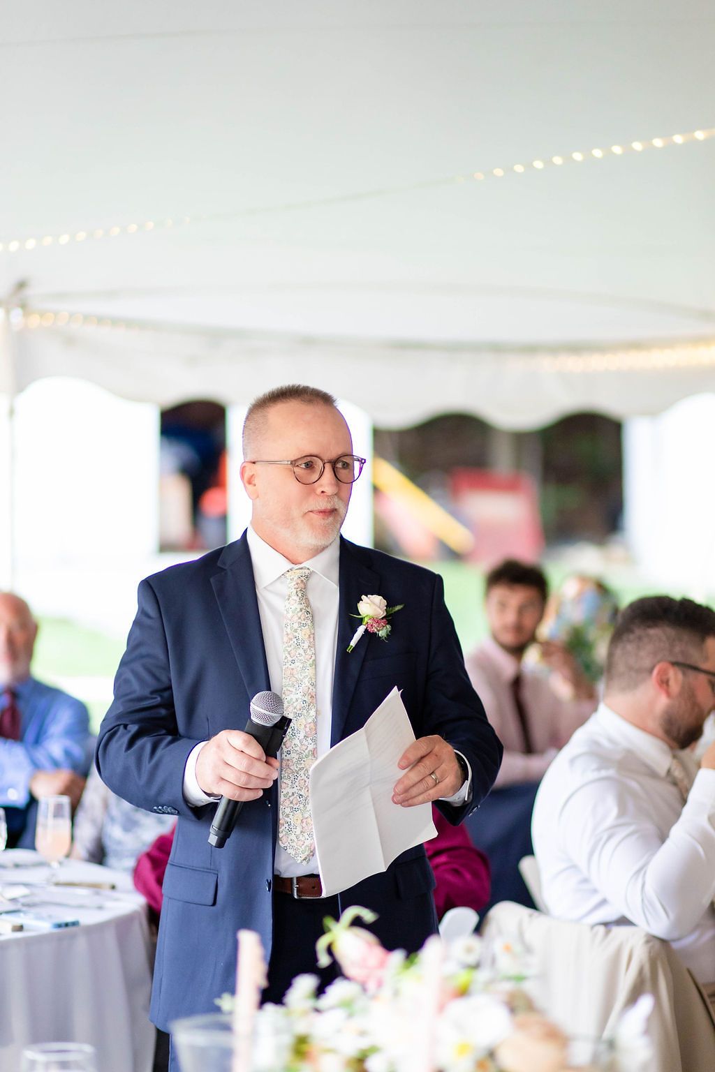 Man in navy suit with boutonniere delivers a wedding speech holding microphone and notes under a tent.
