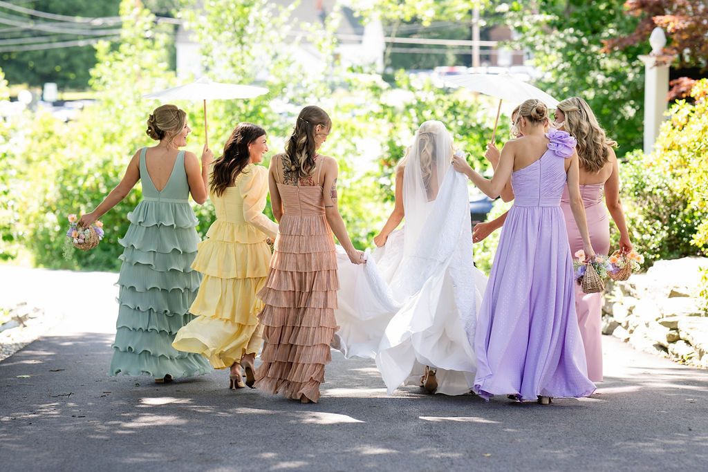 Bride and bridesmaids in pastel rainbow gowns walk hand-in-hand down a sunny garden pathway.