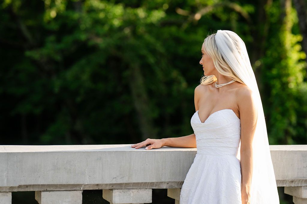 Elegant bride in strapless white gown and veil stands gracefully by stone balustrade outdoors.
