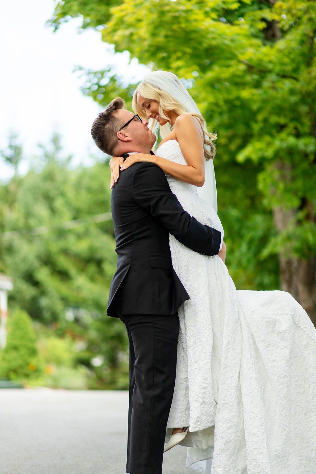 Groom lifting bride in white wedding dress outdoors with lush green trees in background.