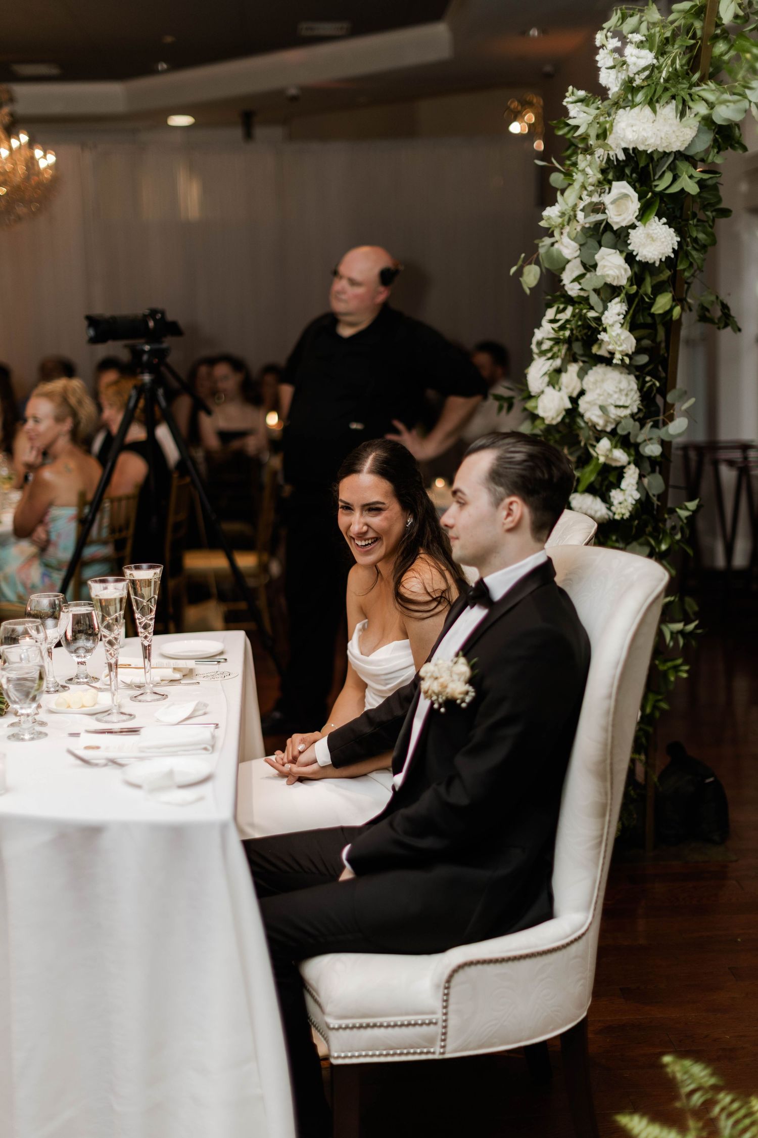 Bride and groom laughing together at sweetheart table adorned with white floral arrangement during wedding reception.