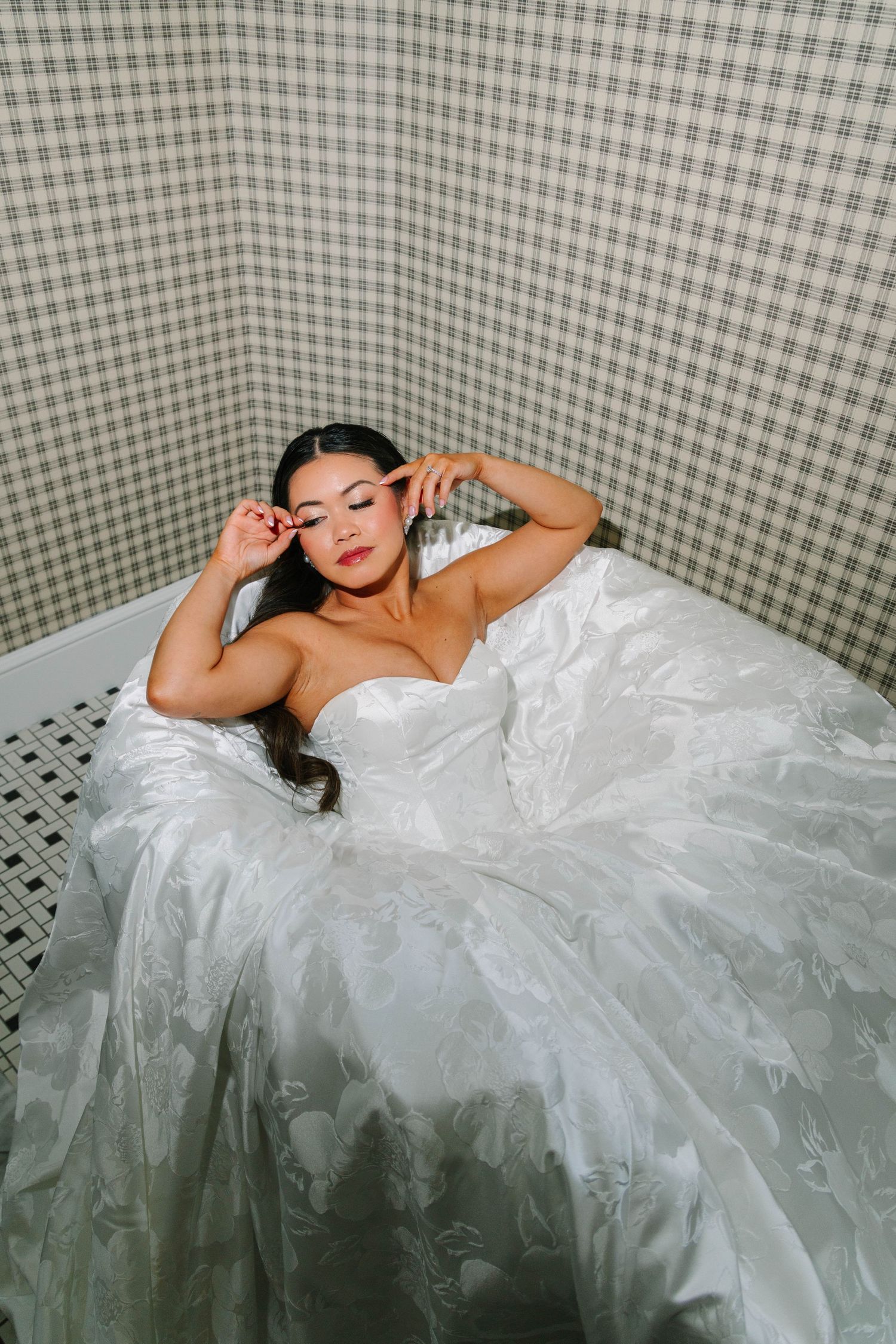 Bride in a voluminous white ballgown reclines in a bathtub, adjusting her makeup before the wedding ceremony.