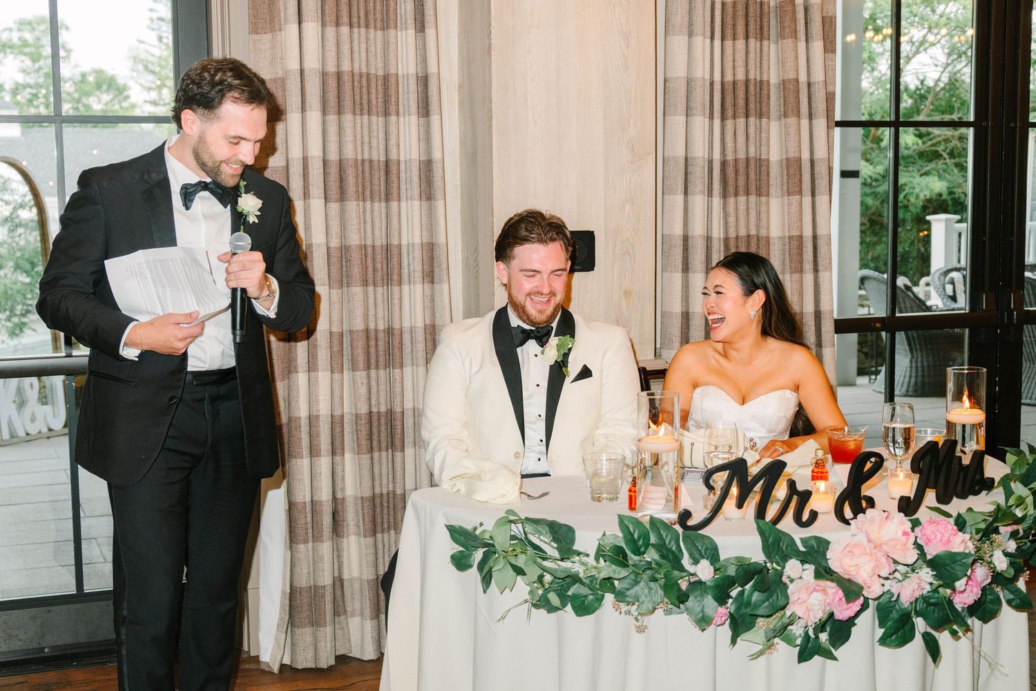 Best man delivers wedding toast as bride and groom smile at decorated sweetheart table.