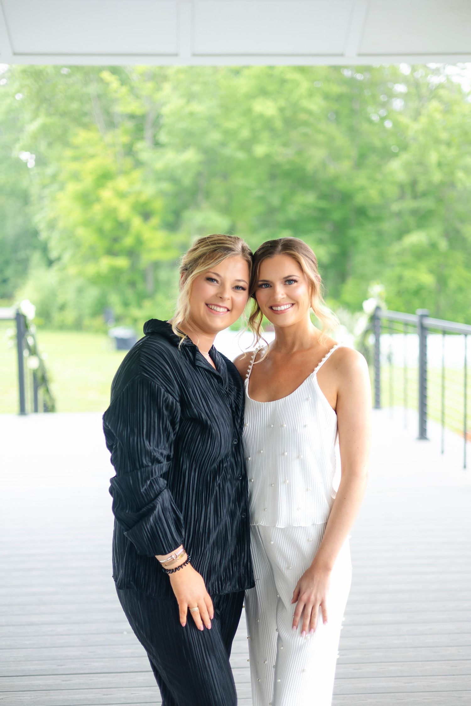 Bride in white pajamas posing with bridesmaid in black pinstripe suit on outdoor deck with greenery.