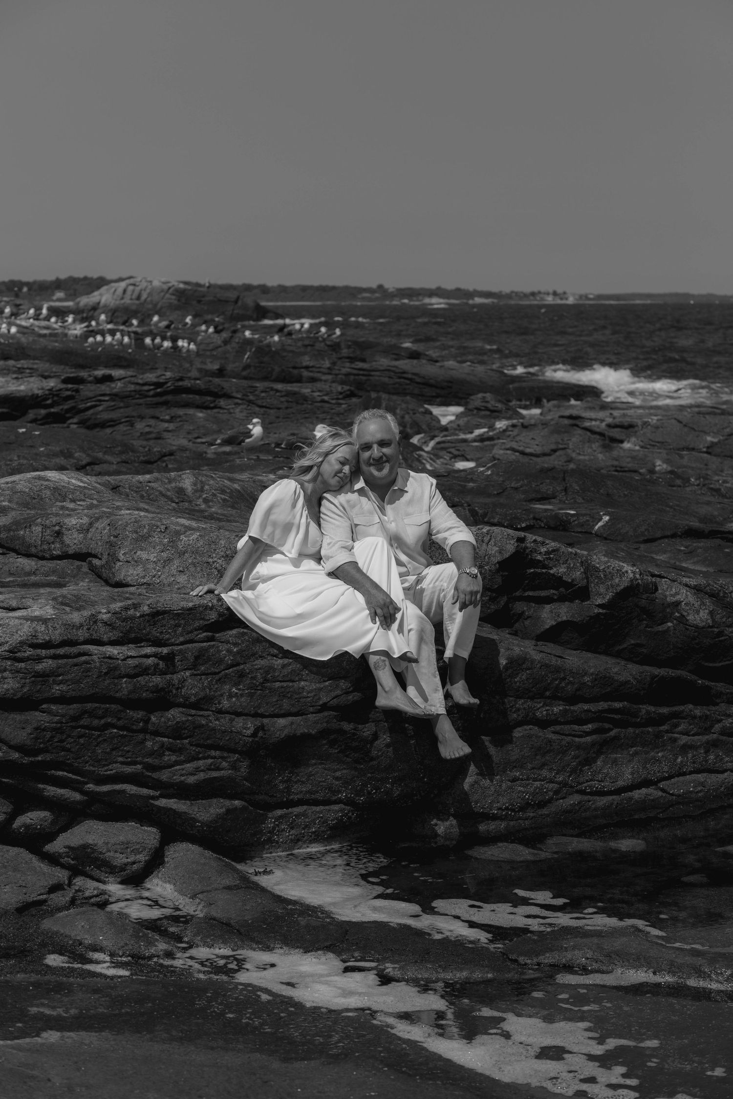 Black and white photo of a couple sitting together on coastal rocks by the ocean, dressed in white outfits.