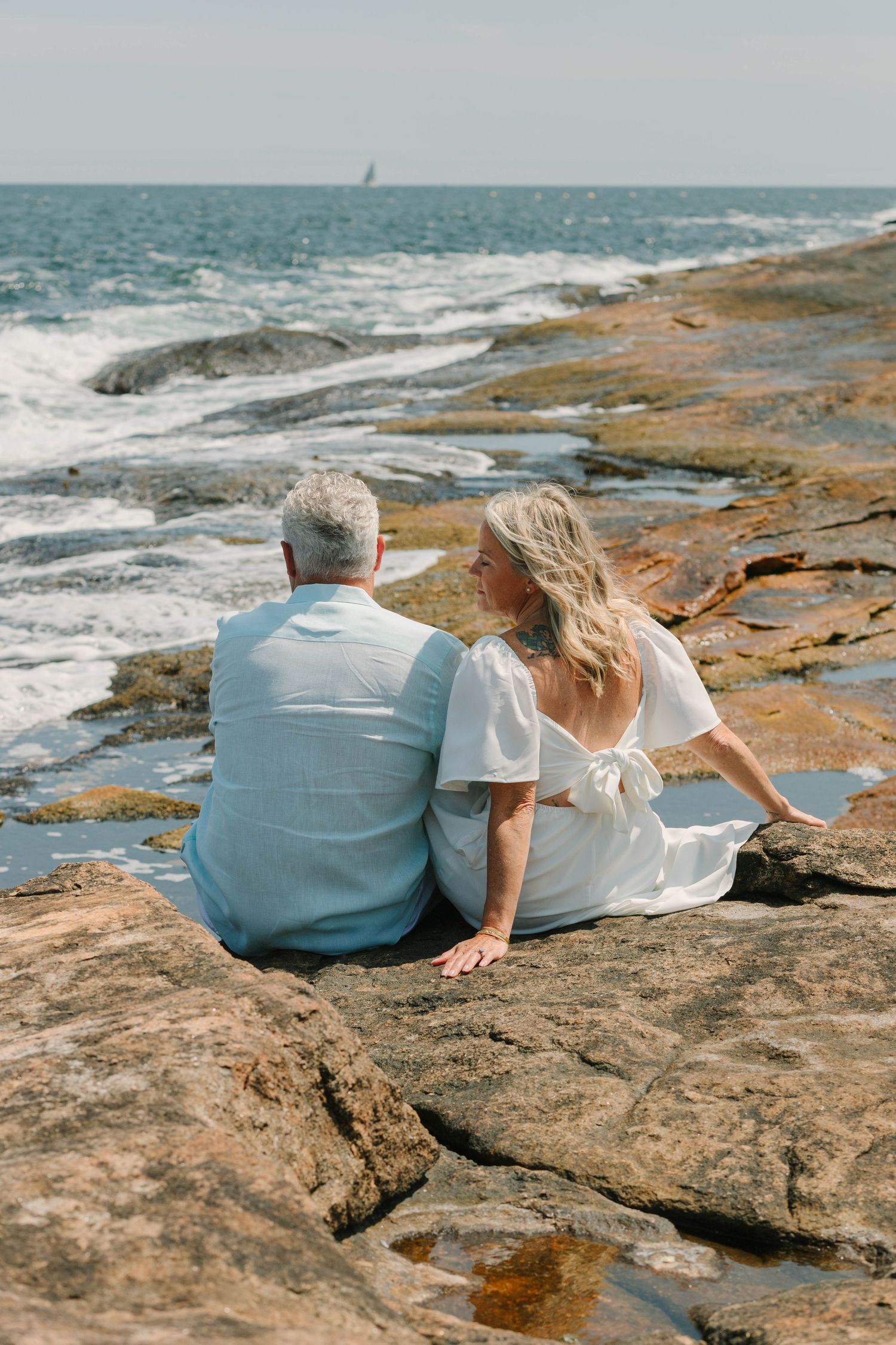 Couple sitting together on rocky oceanside cliff watching waves roll in on a bright sunny day.