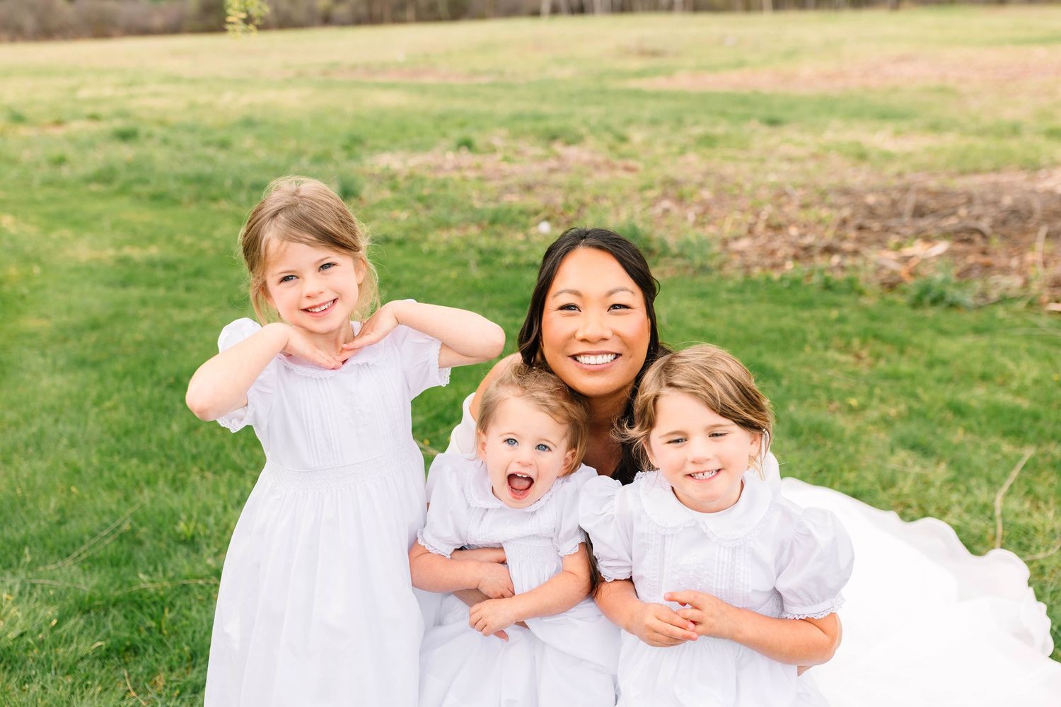 A smiling Asian woman posing outdoors with three young girls in white dresses on green grass.