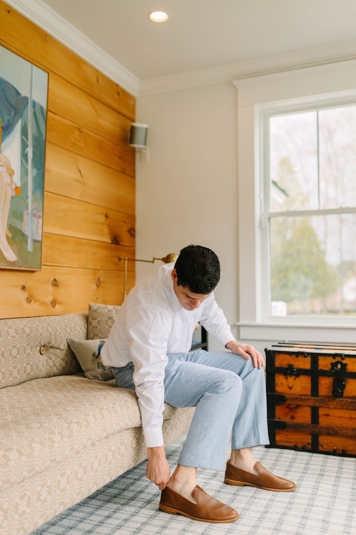 Man in white shirt and light blue pants putting on brown loafers while seated on a beige sofa indoors.