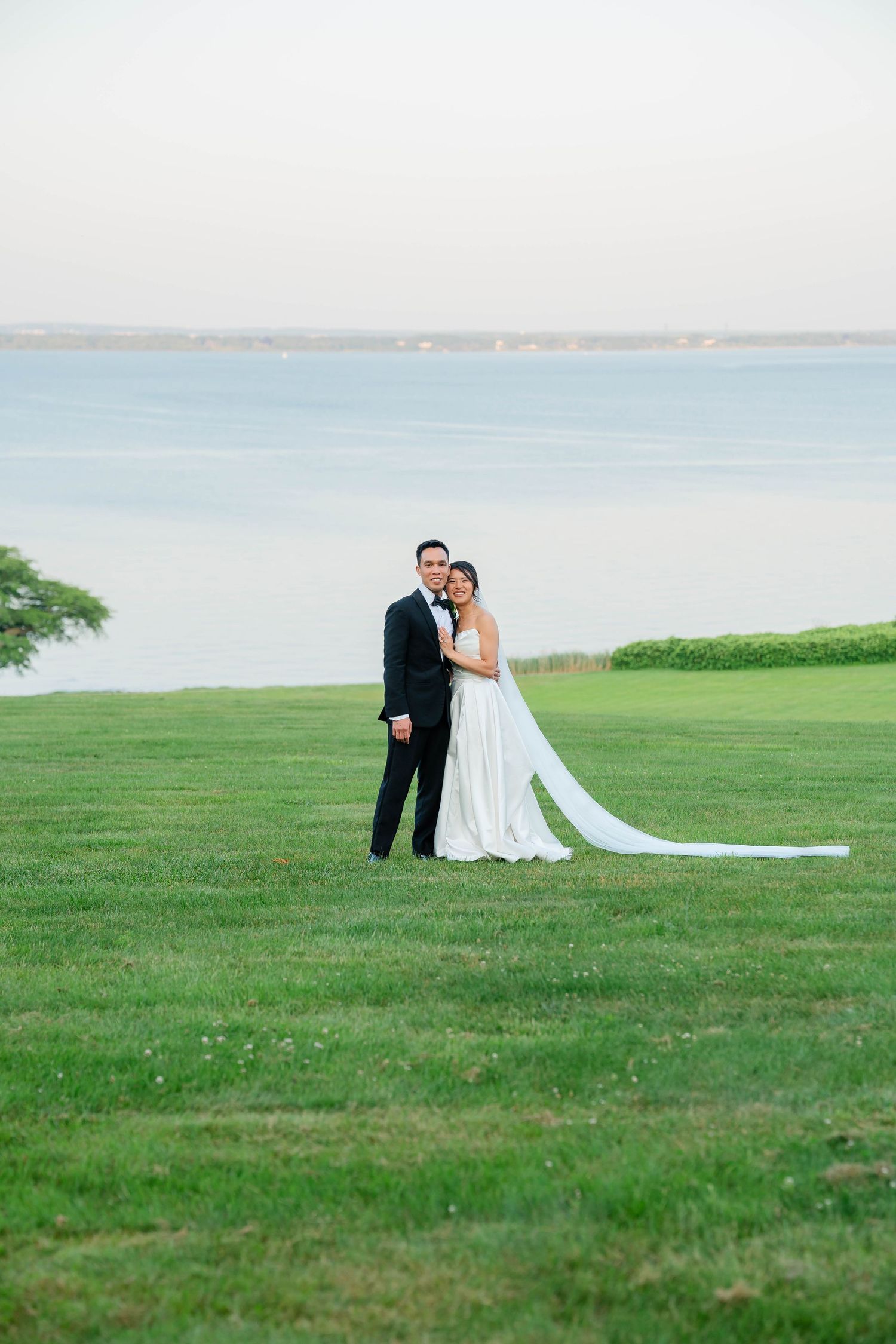 Bride and groom posing on lush green lawn with serene waterfront backdrop at dusk.