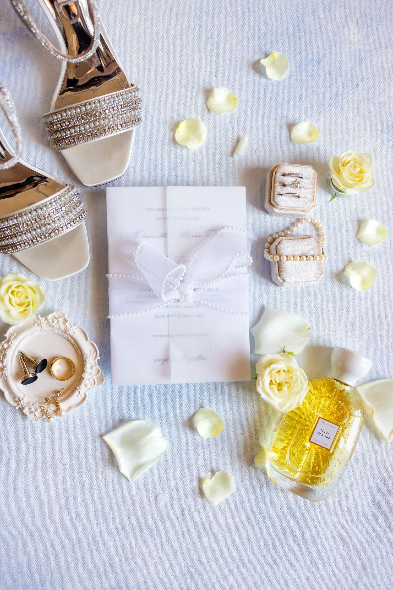 Elegant wedding flat lay with bridal heels, invitation, rings, perfume, and white roses on blue background.