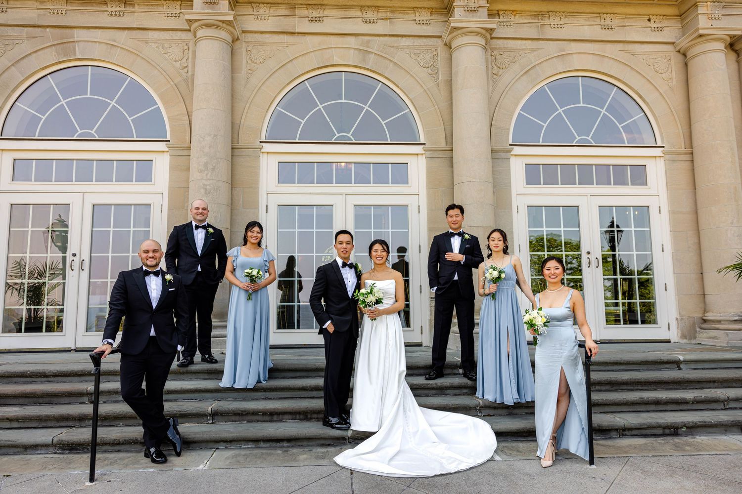 Wedding party poses on elegant venue steps, bride in white gown with bridesmaids in blue dresses and groomsmen in black tuxedos.