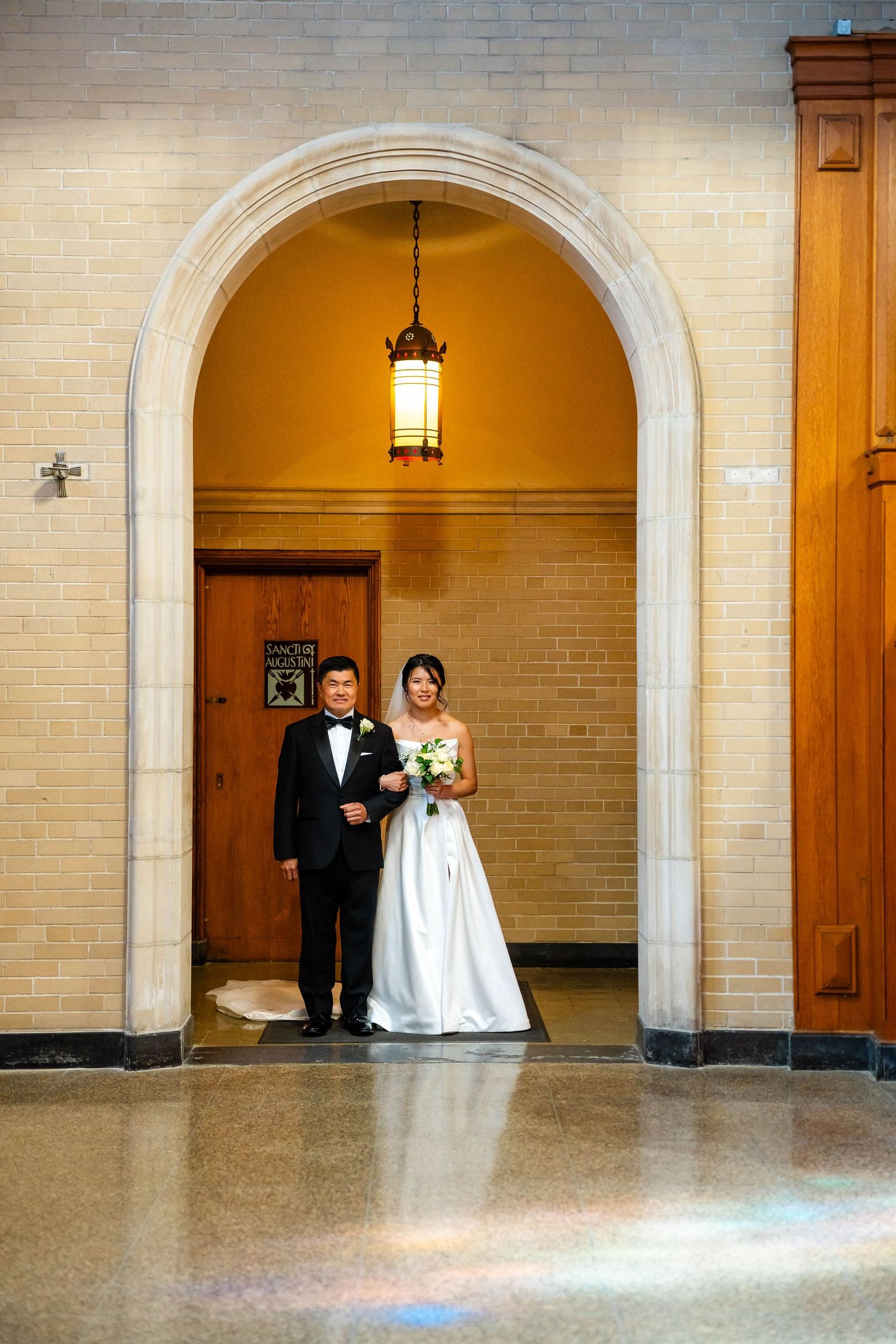 Bride and groom stand together under stone arch with pendant light, bride holding white bouquet.