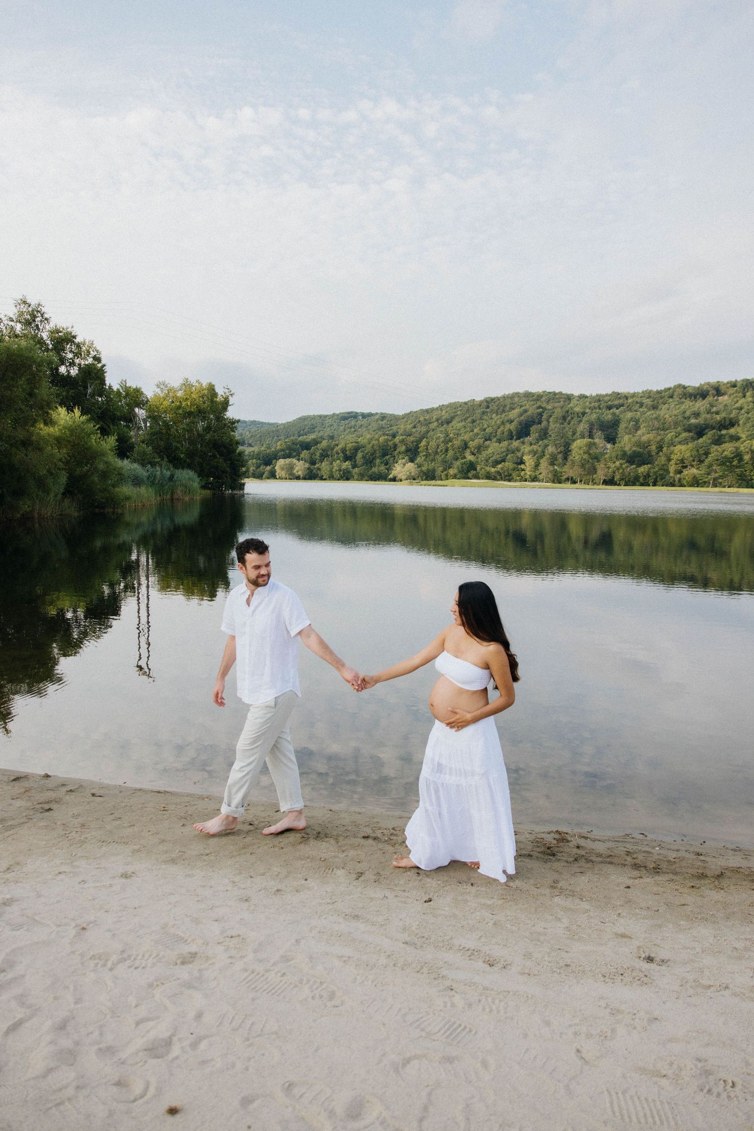Pregnant couple in white outfits holding hands on sandy lakeside beach with lush green forest reflecting in calm water.