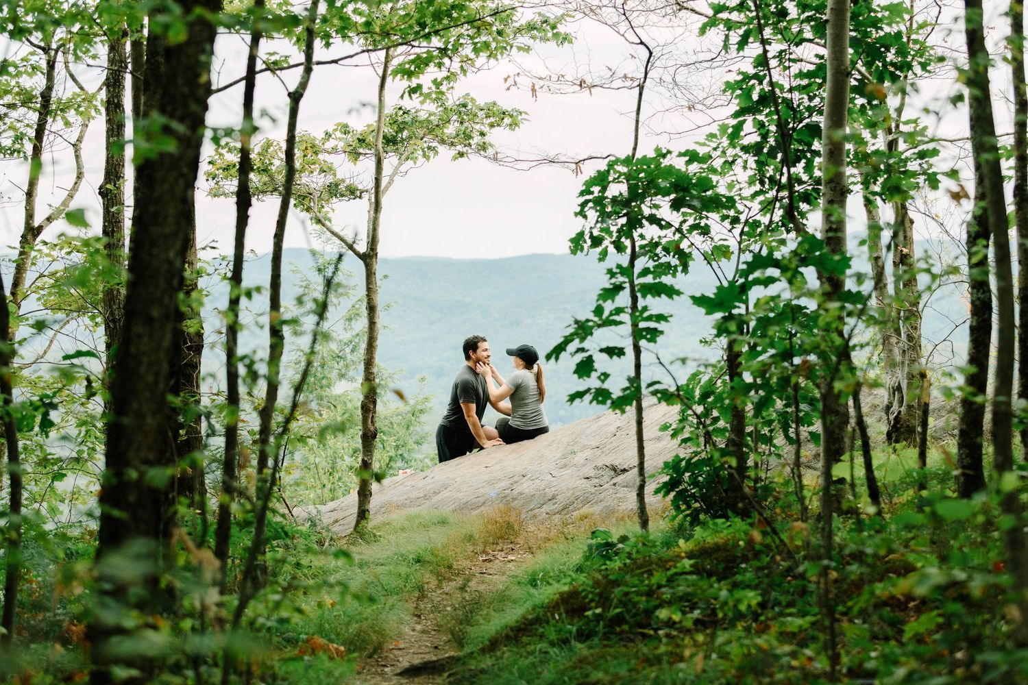 Two people sitting together on a rocky outcrop framed by green trees with scenic mountain landscape behind them.