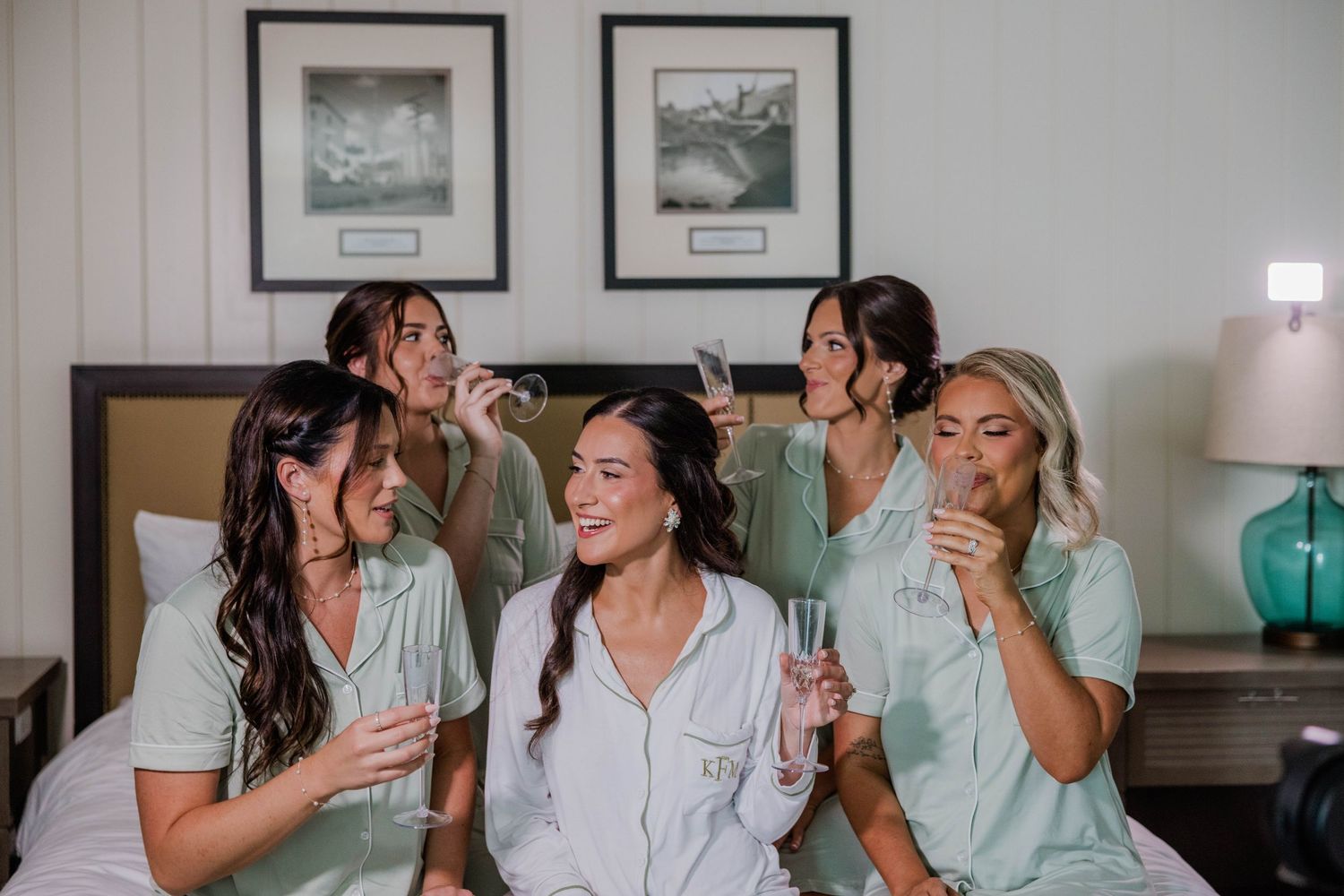 Bridesmaids in mint green pajamas sipping champagne with bride in white on a bed during wedding morning preparations.
