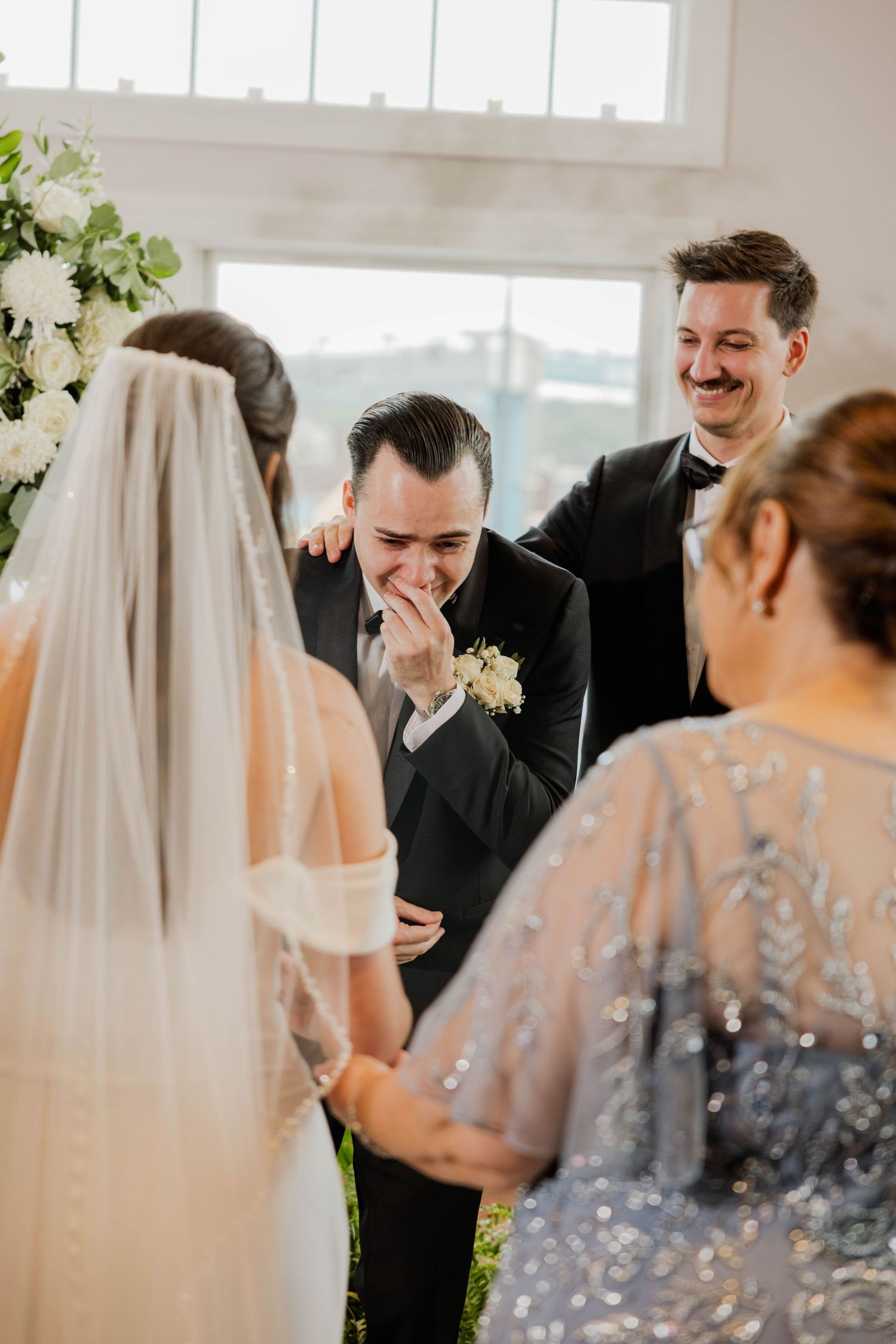 Emotional groom covers his mouth while crying as bride approaches during indoor wedding ceremony.
